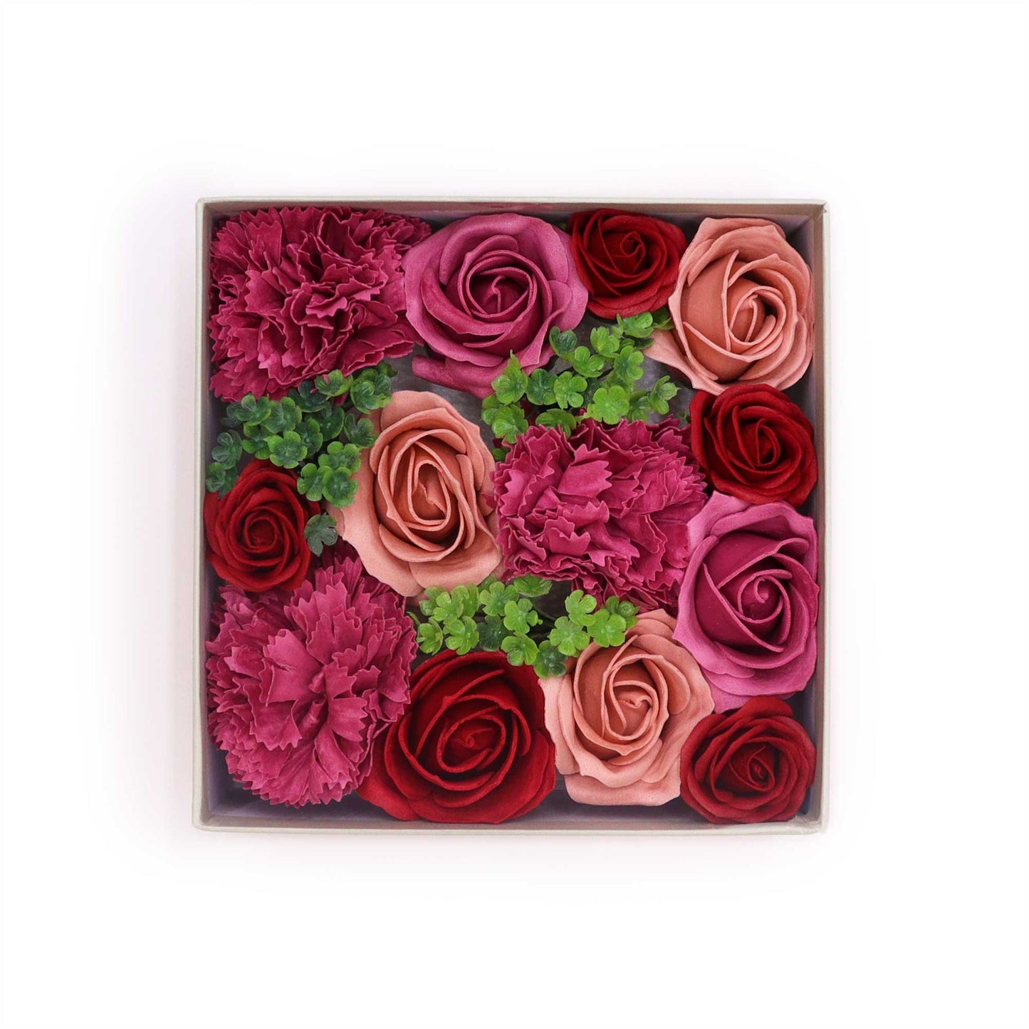 Roses and carnations of various shades of pink and red are arranged in a square box.  The flowers are nestled together with small green leaves.  The box sits on a white background.
