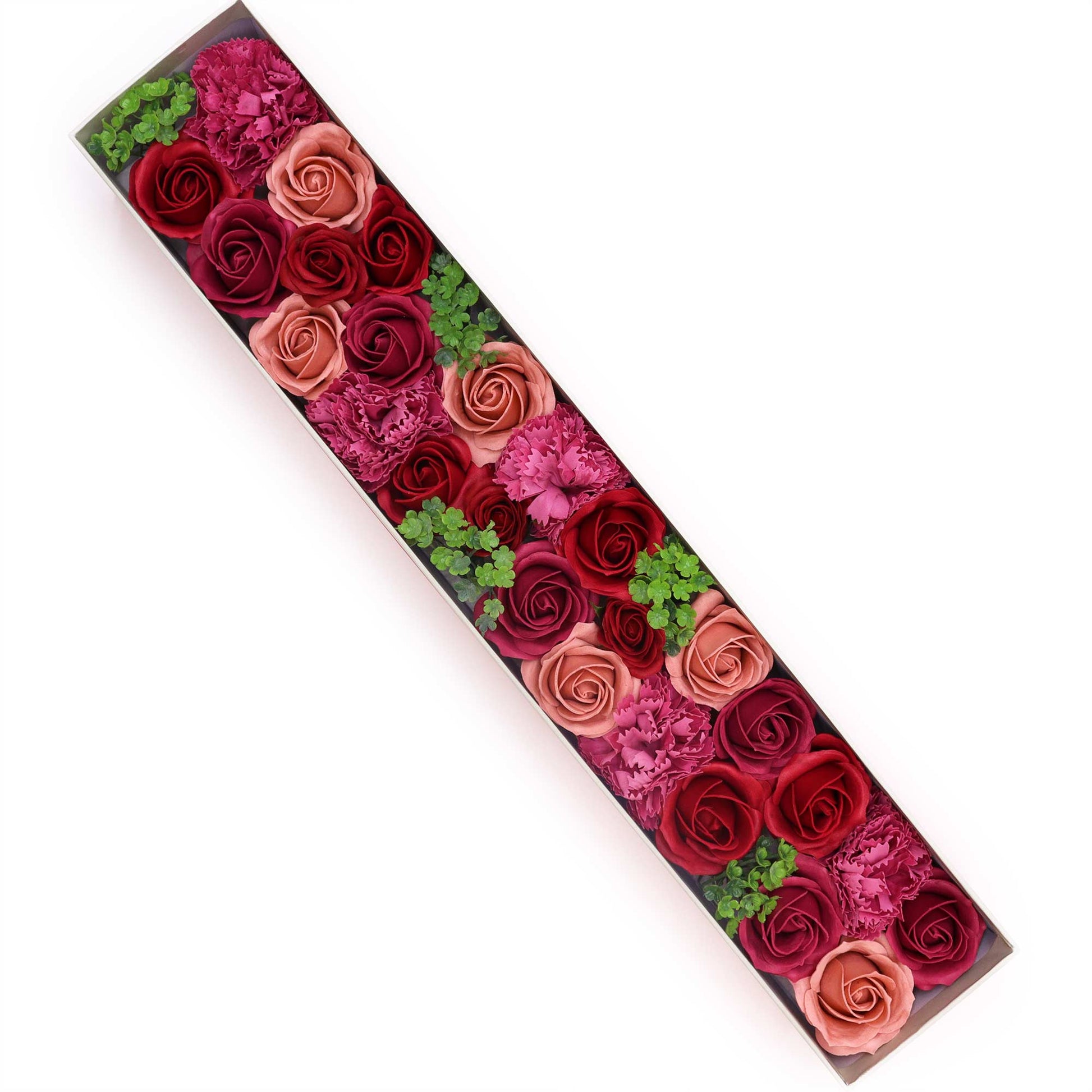 A long, rectangular box contains numerous roses and carnations of varying shades of pink and red, arranged in a diagonal line.  Small green sprigs are interspersed. The box rests on a white background.