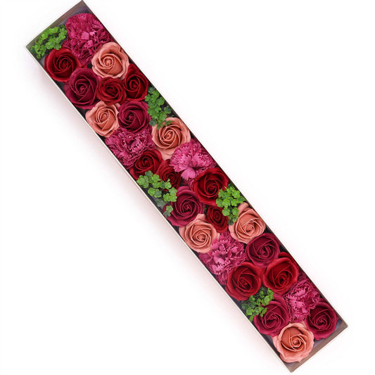 A long, rectangular box contains numerous roses and carnations of varying shades of pink and red, arranged in a diagonal line.  Small green sprigs are interspersed. The box rests on a white background.