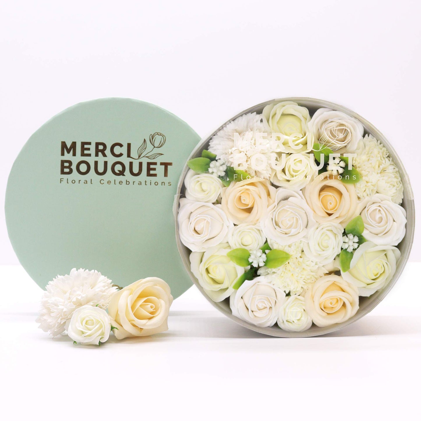 A circular arrangement of cream and white soap roses and other flowers sits inside a gray box.  It rests next to a light green box lid with “MERCI BOUQUET Floral Celebrations” printed on it.  Three additional soap flowers are placed near the box.
