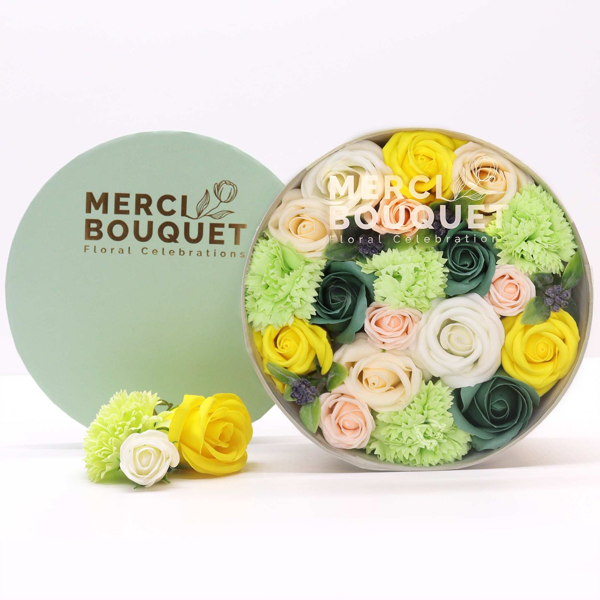 A round box containing a soap flower bouquet of pastel and yellow roses and carnations sits on a white surface.  A smaller arrangement of soap flowers is nearby. The box says: MERCI BOUQUET Floral Celebrations.  The lid also says MERCI BOUQUET Floral Celebrations.