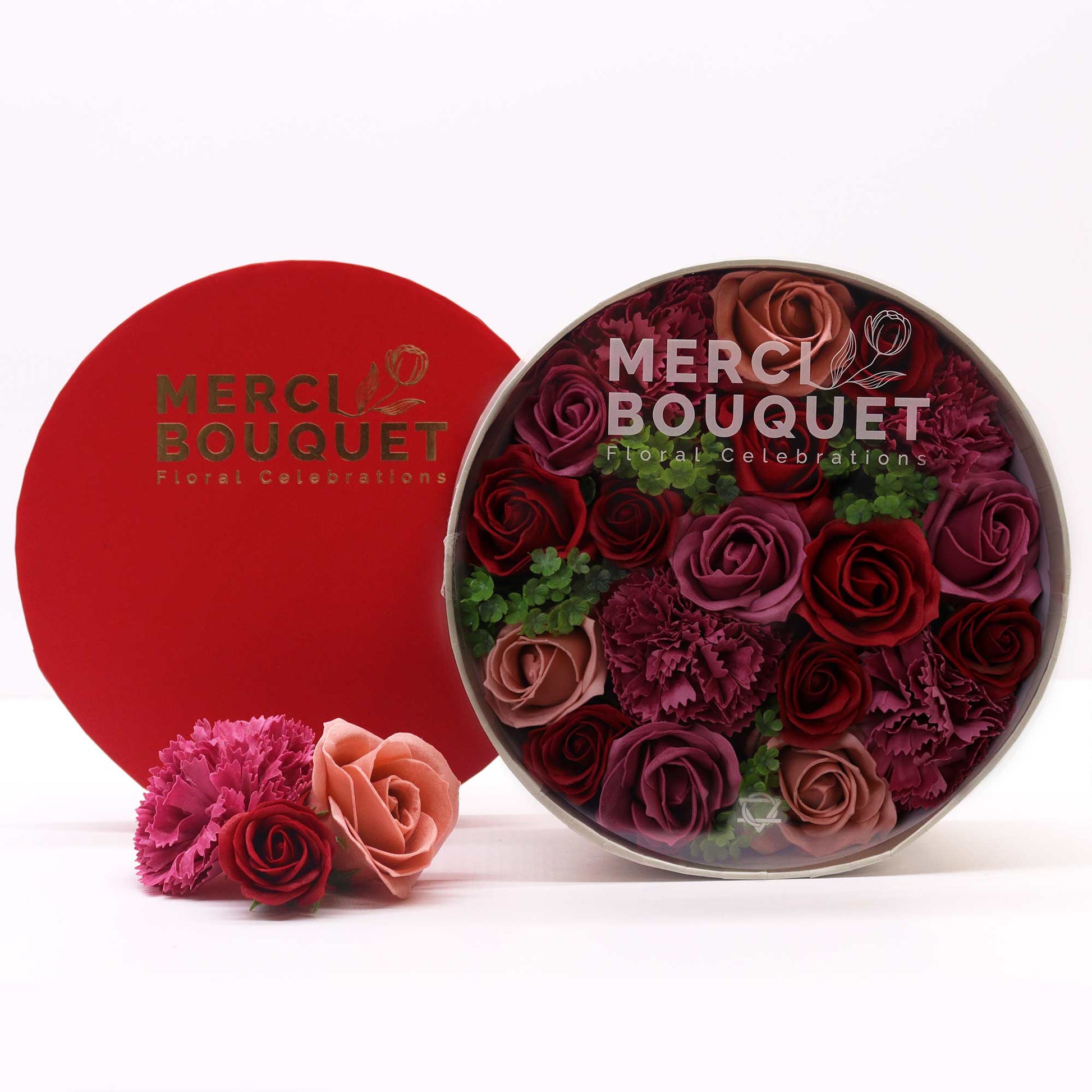 A round box containing a bouquet of preserved roses and carnations in varying shades of red and pink sits on a white surface next to a red box.  The boxes are labeled “MERCI BOUQUET Floral Celebrations.”
