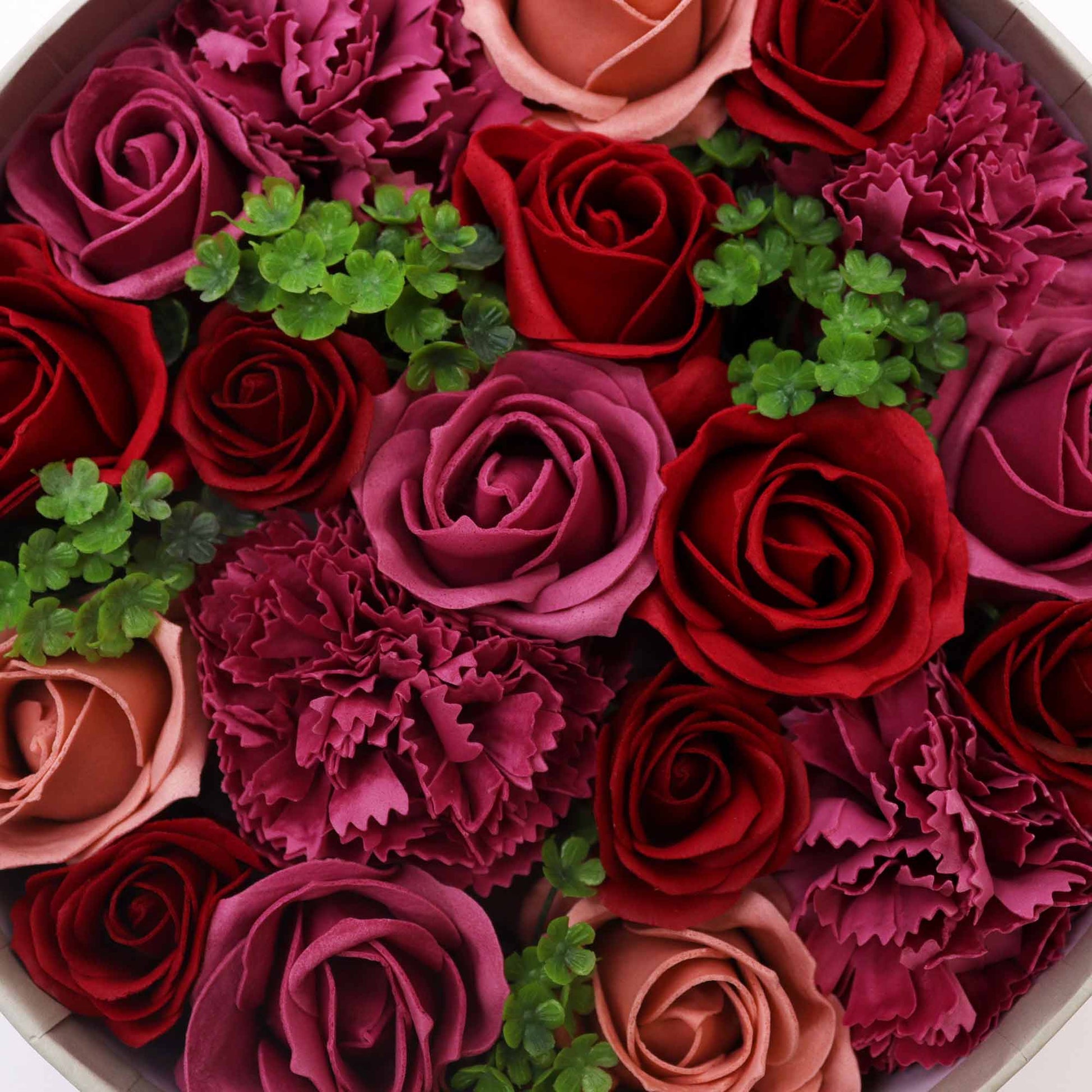 Soap roses and carnations, arranged in a circular box, are densely packed together with small green filler plants.