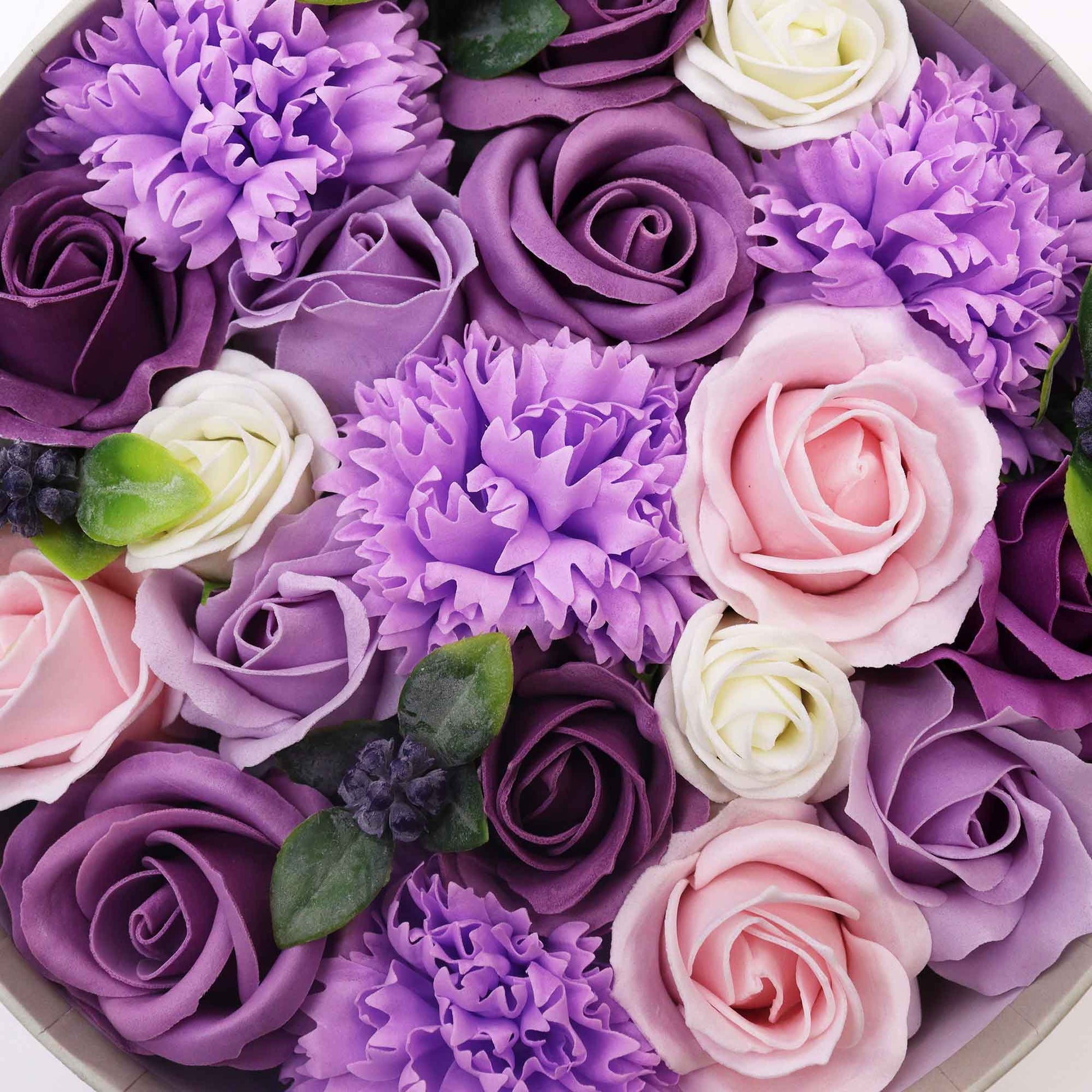 Soap flowers, arranged in a circular box, comprise various shades of purple, pink, and white roses and carnations.
