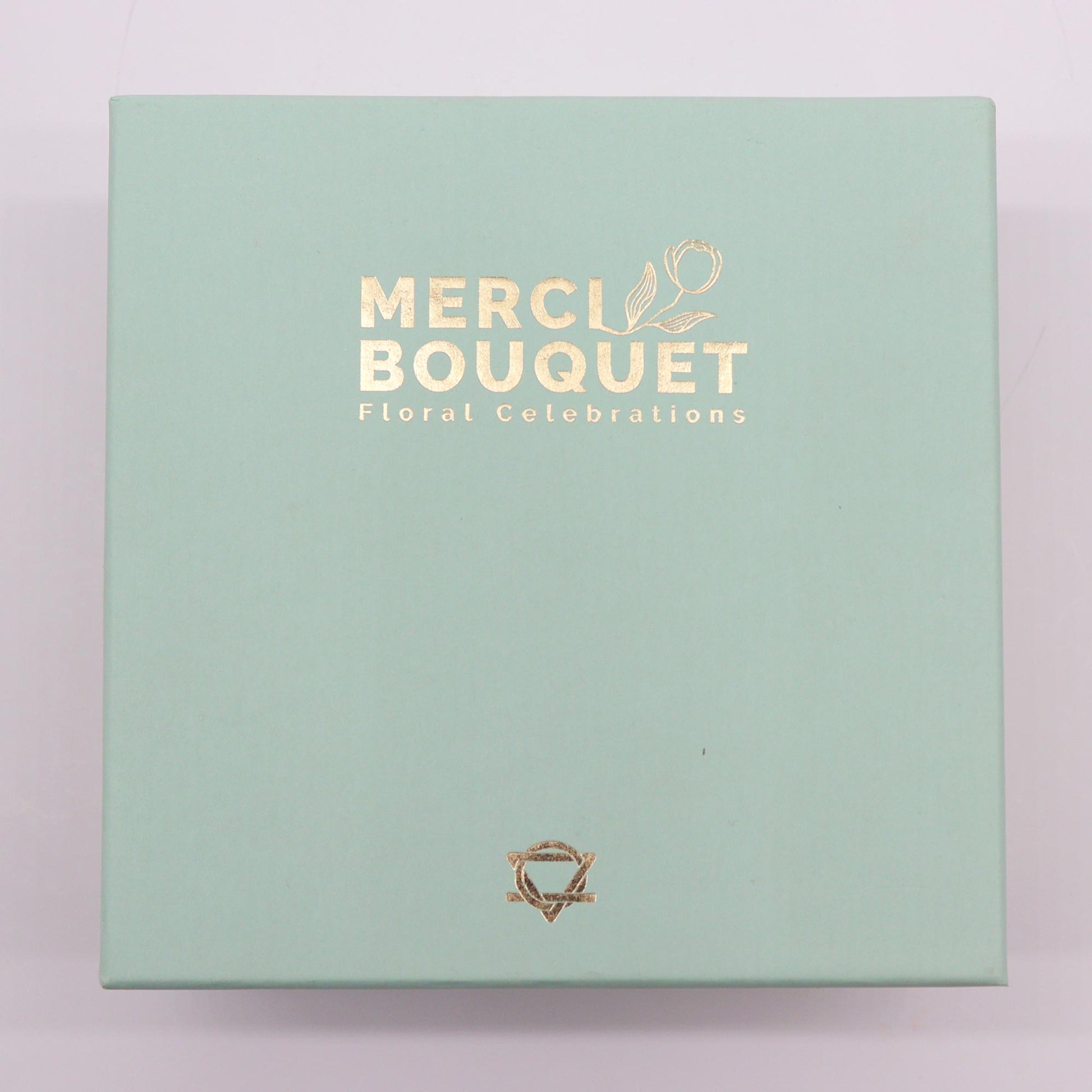 A light-green box rests on a surface.  It displays gold lettering: "MERCI BOUQUET Floral Celebrations" and a small gold logo.
