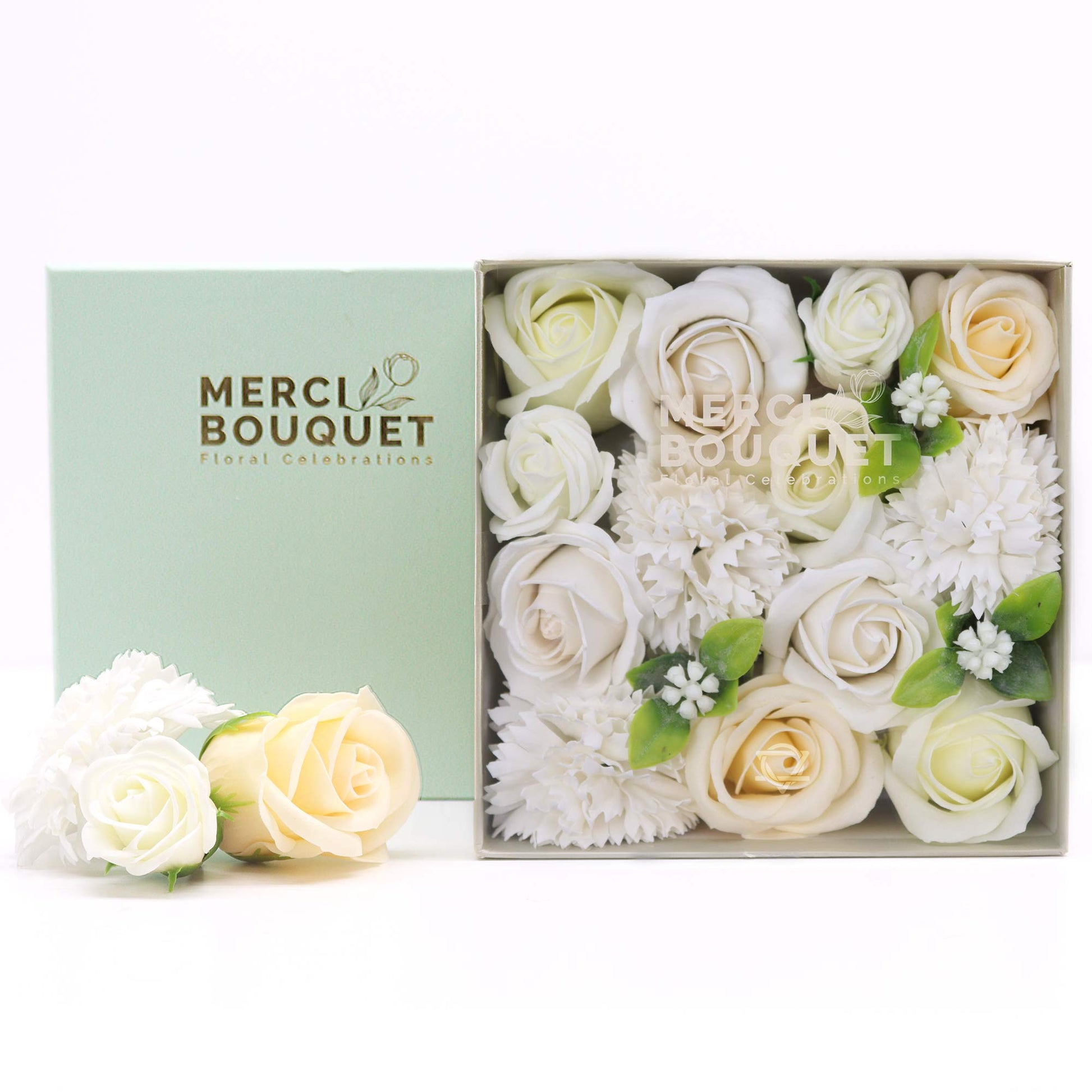 A square box contains an arrangement of off-white and cream soap roses and carnations.  The box sits on a white surface beside a pale green box that reads: MERCI BOUQUET Floral Celebrations.