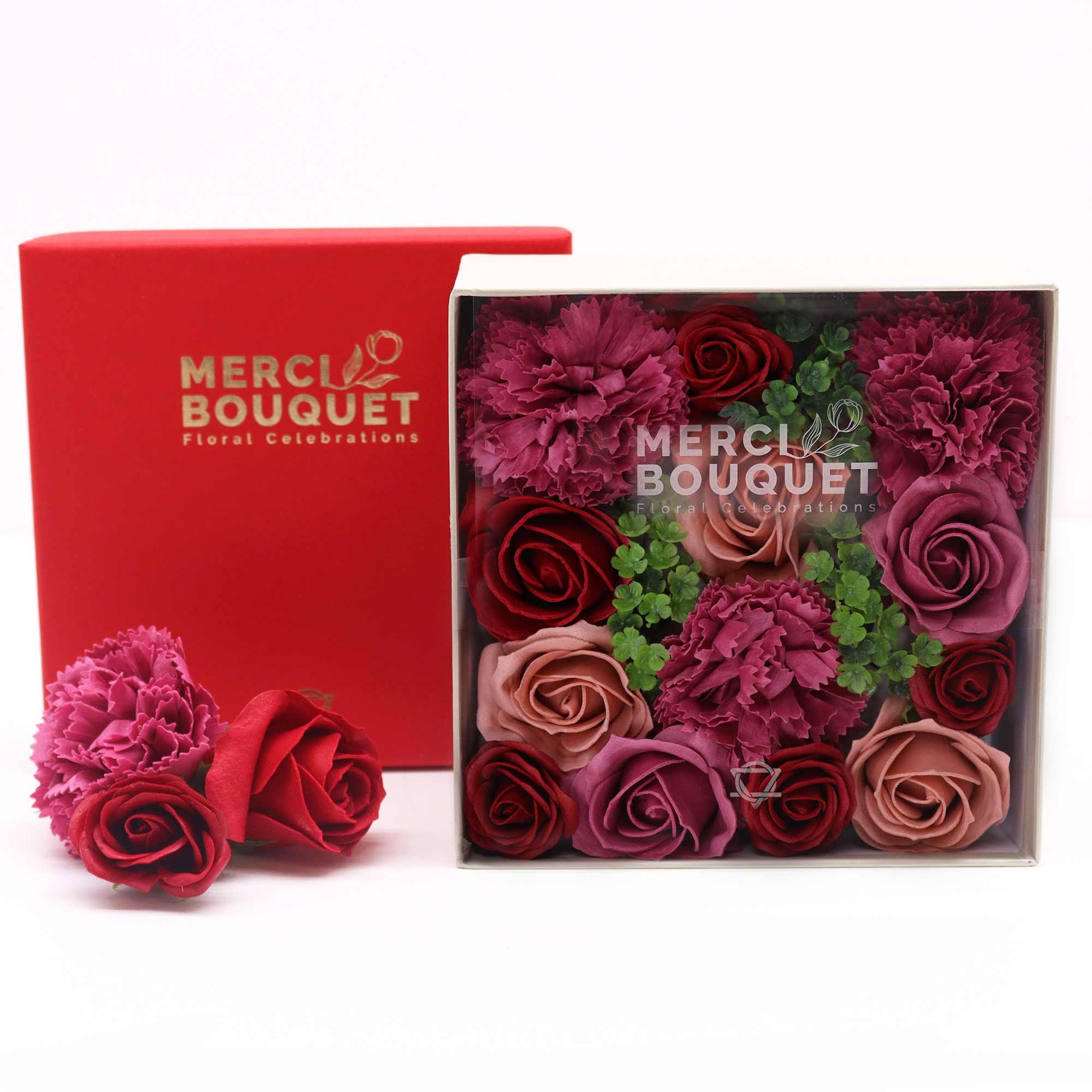 A square box containing an arrangement of soap roses and carnations in various shades of red and pink sits on a white surface next to a red box.  "MERCI BOUQUET Floral Celebrations" is printed on both boxes.