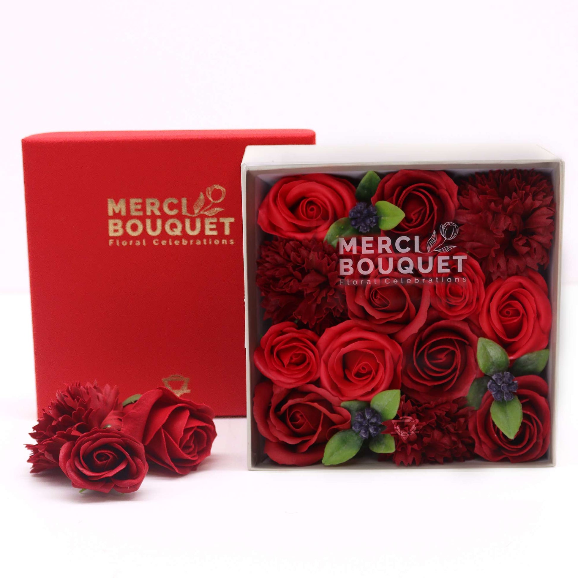 A square box contains red roses and carnations; it sits on a white surface next to a red box.  Both boxes display “MERCI BOUQUET Floral Celebrations.”