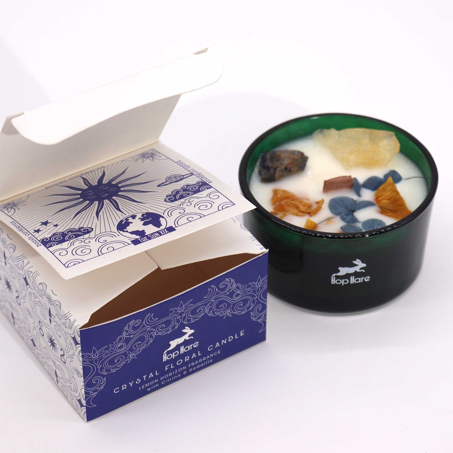 A dark green candle sits beside its opened box on a white surface. The candle has crystals embedded in the wax. The box reads "Hop Hare CRYSTAL FLORAL CANDLE LEMON HORIZON FRAGRANCE with Citrine & Sodalite" and has illustrations including "THE SUN XIX boosts confidence".