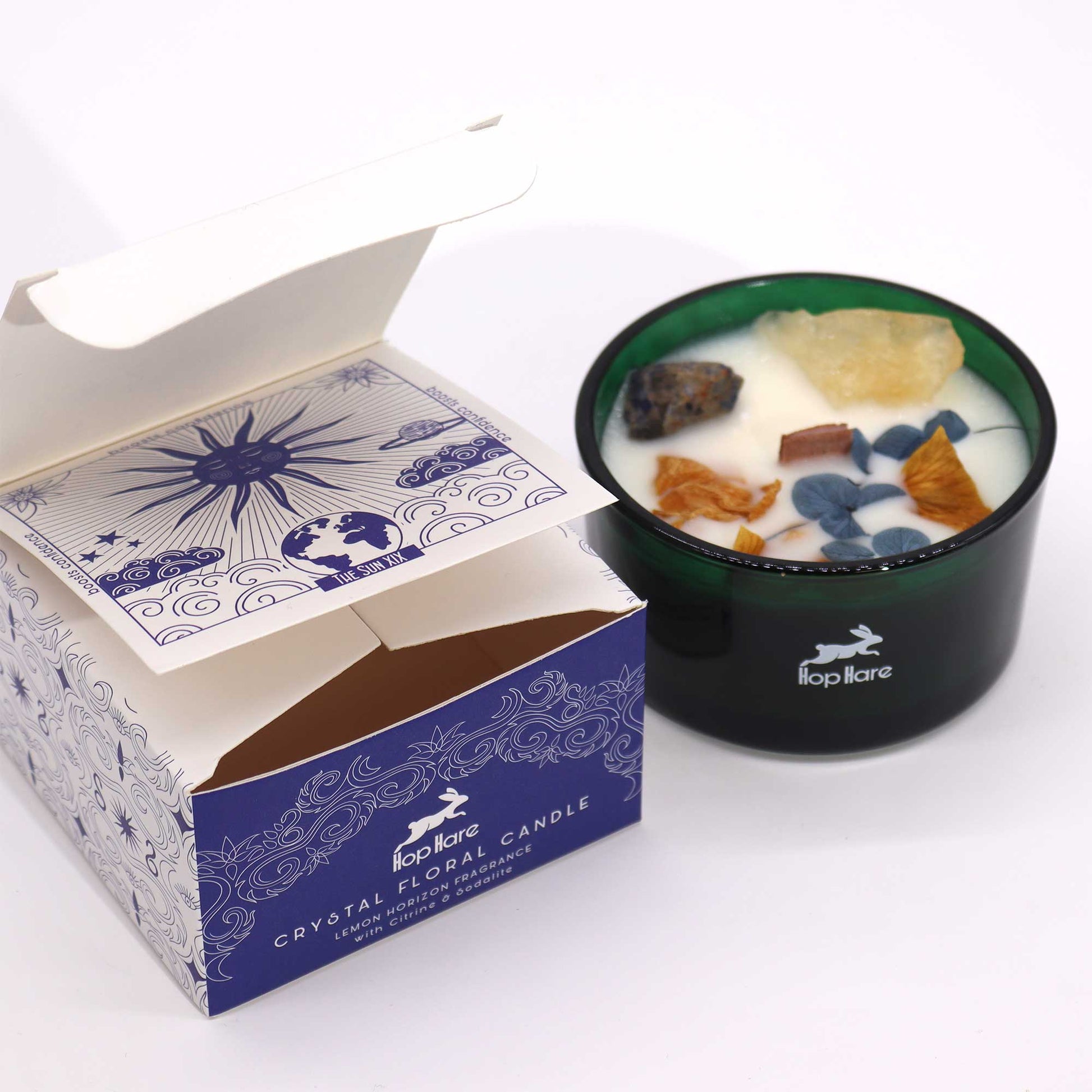 A dark green candle sits beside its opened box on a white surface. The candle has crystals embedded in the wax. The box reads "Hop Hare CRYSTAL FLORAL CANDLE LEMON HORIZON FRAGRANCE with Citrine & Sodalite" and has illustrations including "THE SUN XIX boosts confidence".