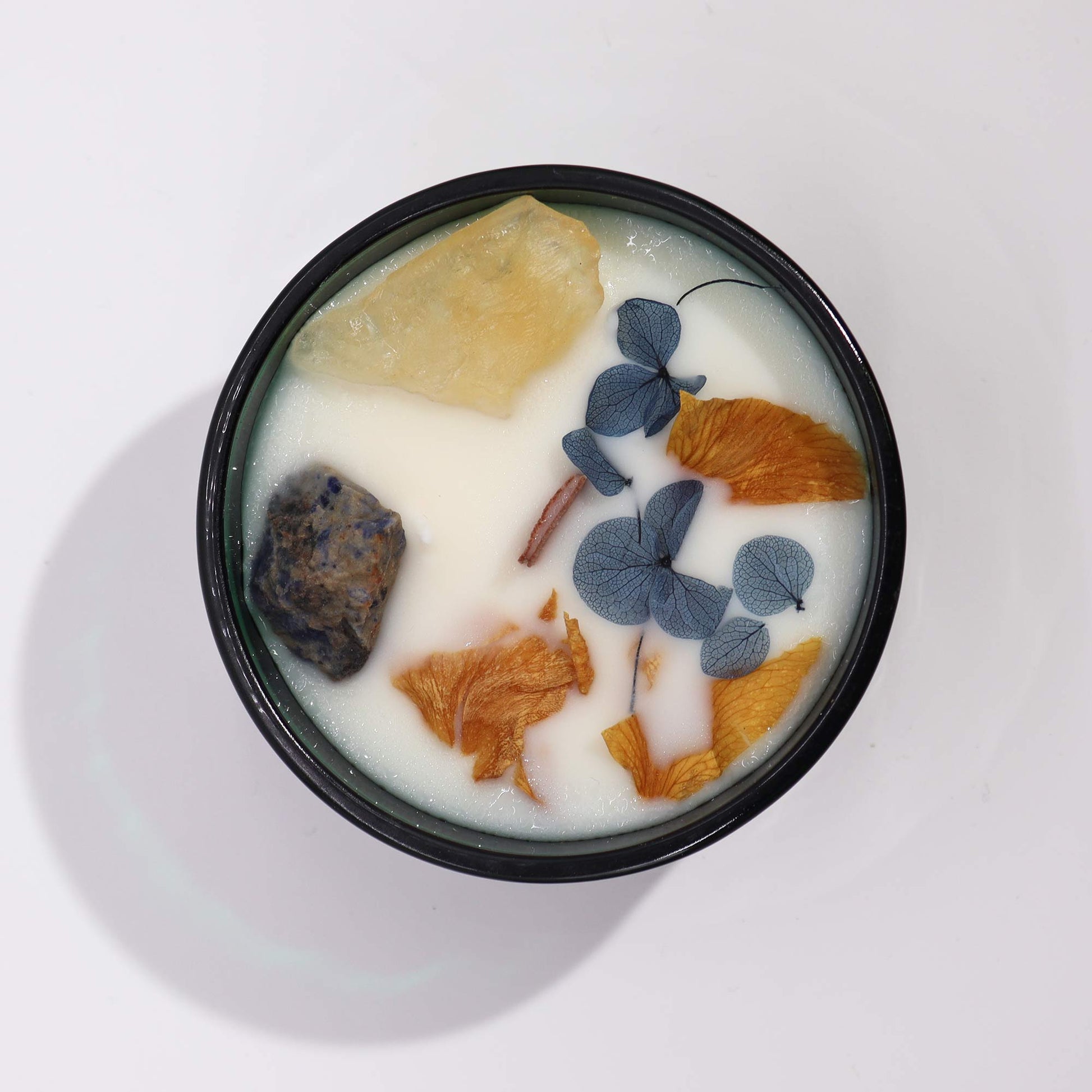 A candle sits flat against a white backdrop. It is in a black container, and a waxy white substance with gems and dried flowers is inside the container.