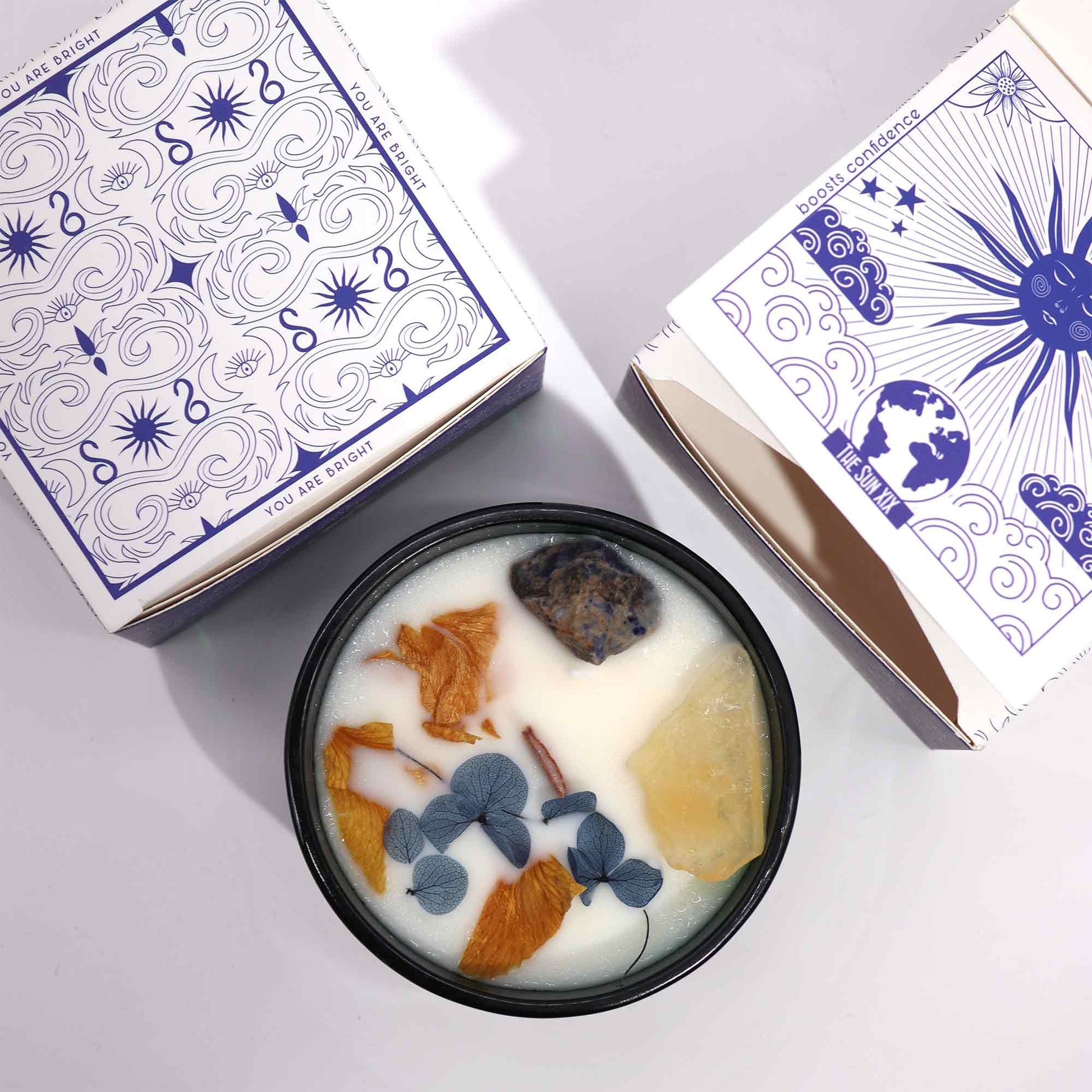 A candle sits in a black tin, adorned with crystals and foliage, nestled between its packaging. Two boxes are seen: One features the words "YOU ARE BRIGHT", while the other reads "THE SUN XIX" and "boosts confidence."