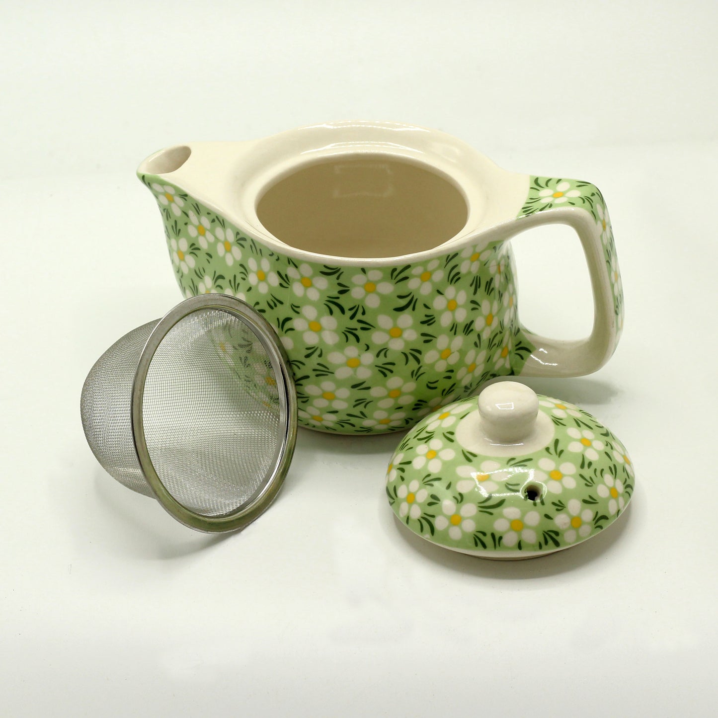 A green and white floral teapot sits with its lid and a metal infuser nearby on a white surface.