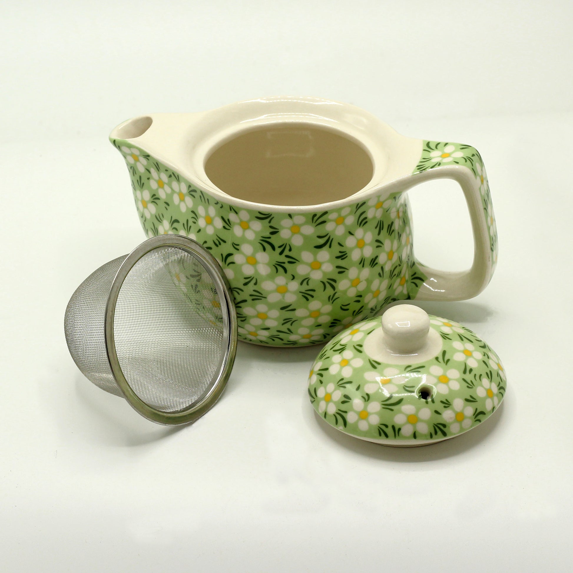 A green and white floral teapot sits with its lid and a metal infuser nearby on a white surface.