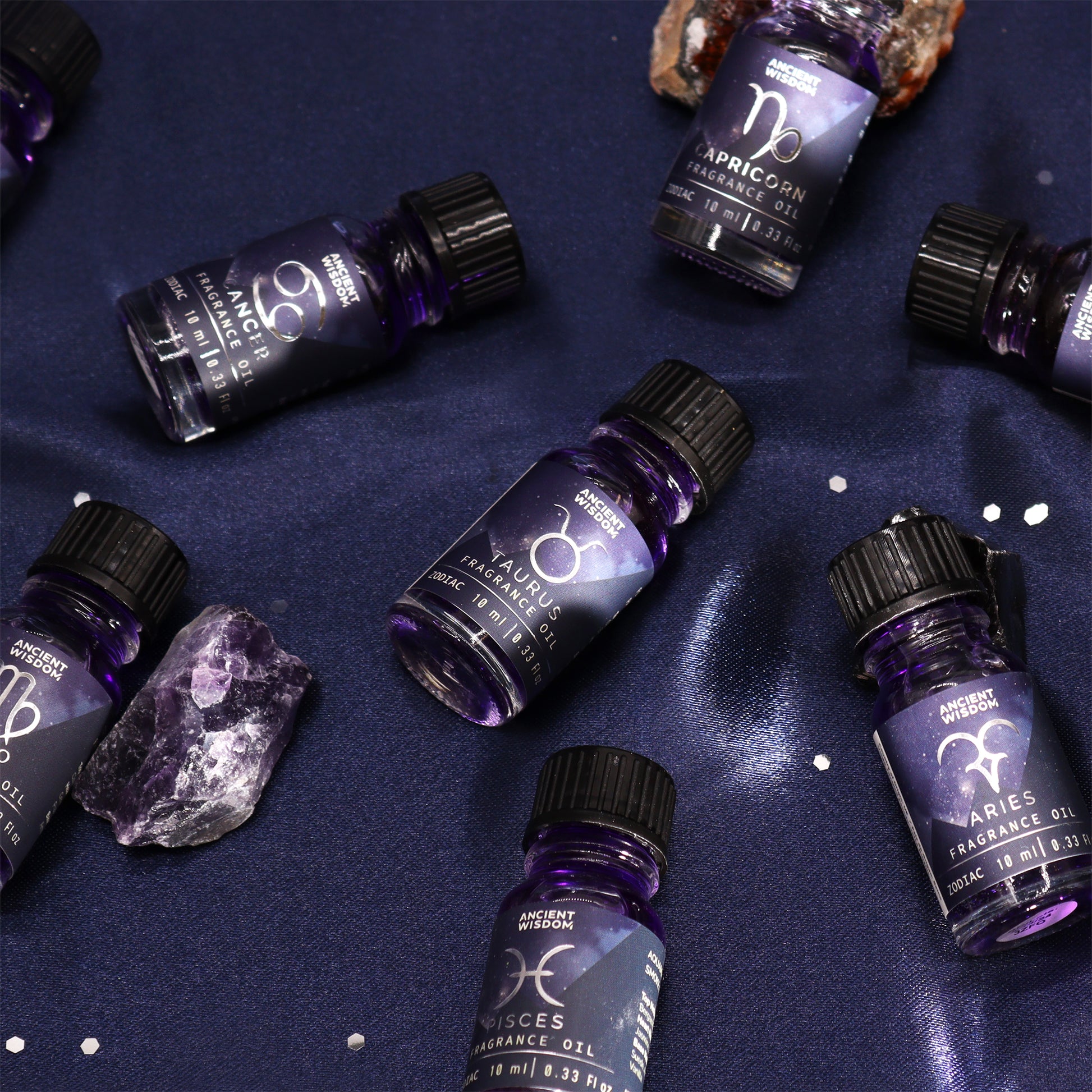 Zodiac fragrance oils rest on a dark blue fabric.  They are arranged amongst small crystals. Each bottle displays its corresponding astrological sign.