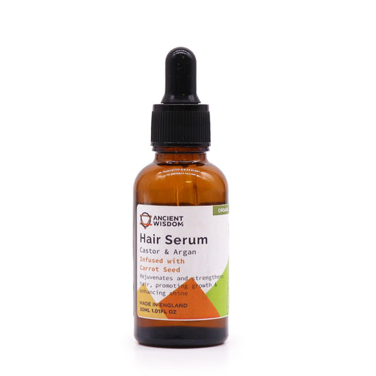 A small amber bottle of "Ancient Wisdom Hair Serum" rests on a white surface. It has a black dropper cap. The label describes Castor & Argan oil infused with Carrot Seed. "MADE IN ENGLAND, 30ML 1.01FL OZ."