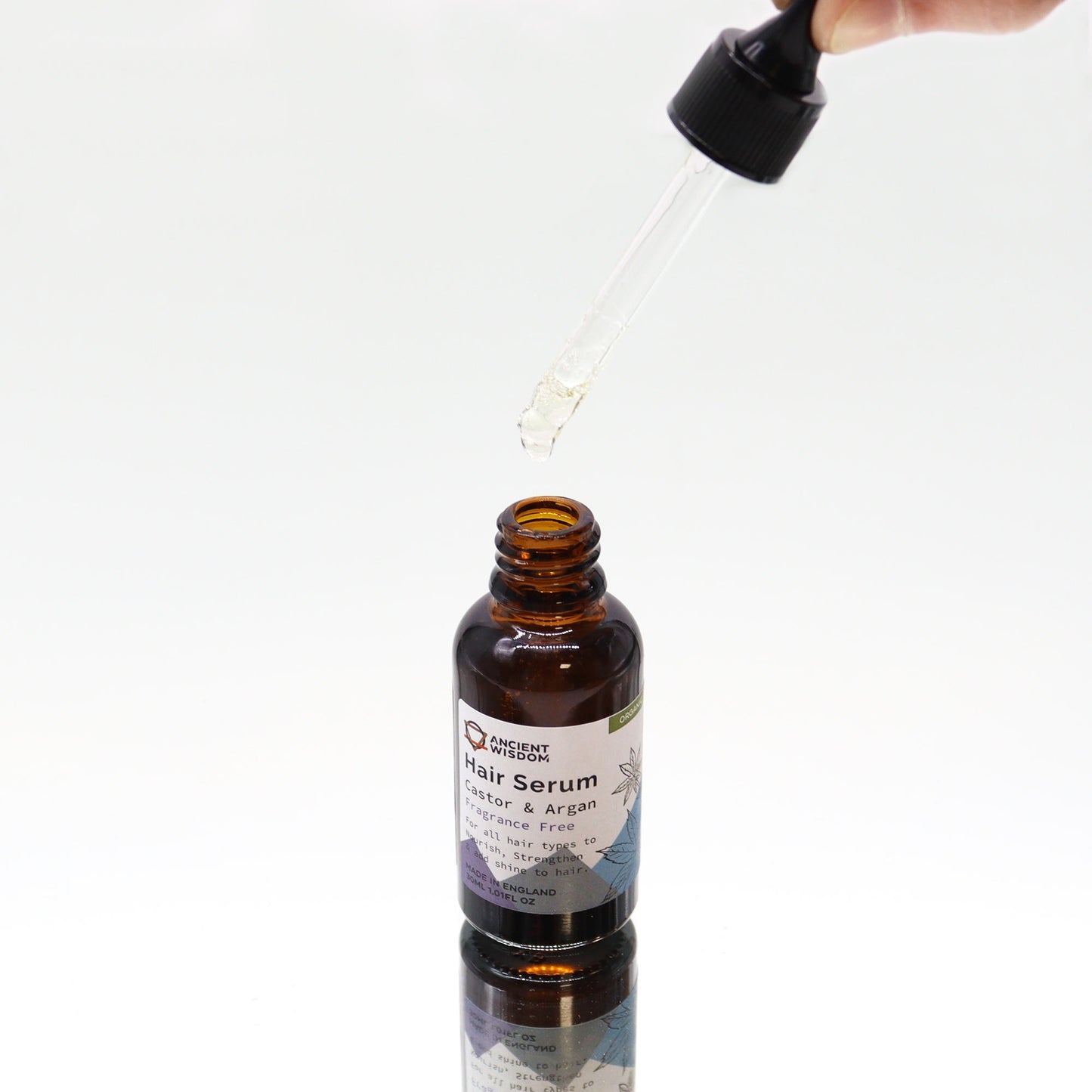 A bottle of "Ancient Wisdom Hair Serum, Castor & Argan, Fragrance Free" is having liquid dispensed from a dropper above it, against a white background.