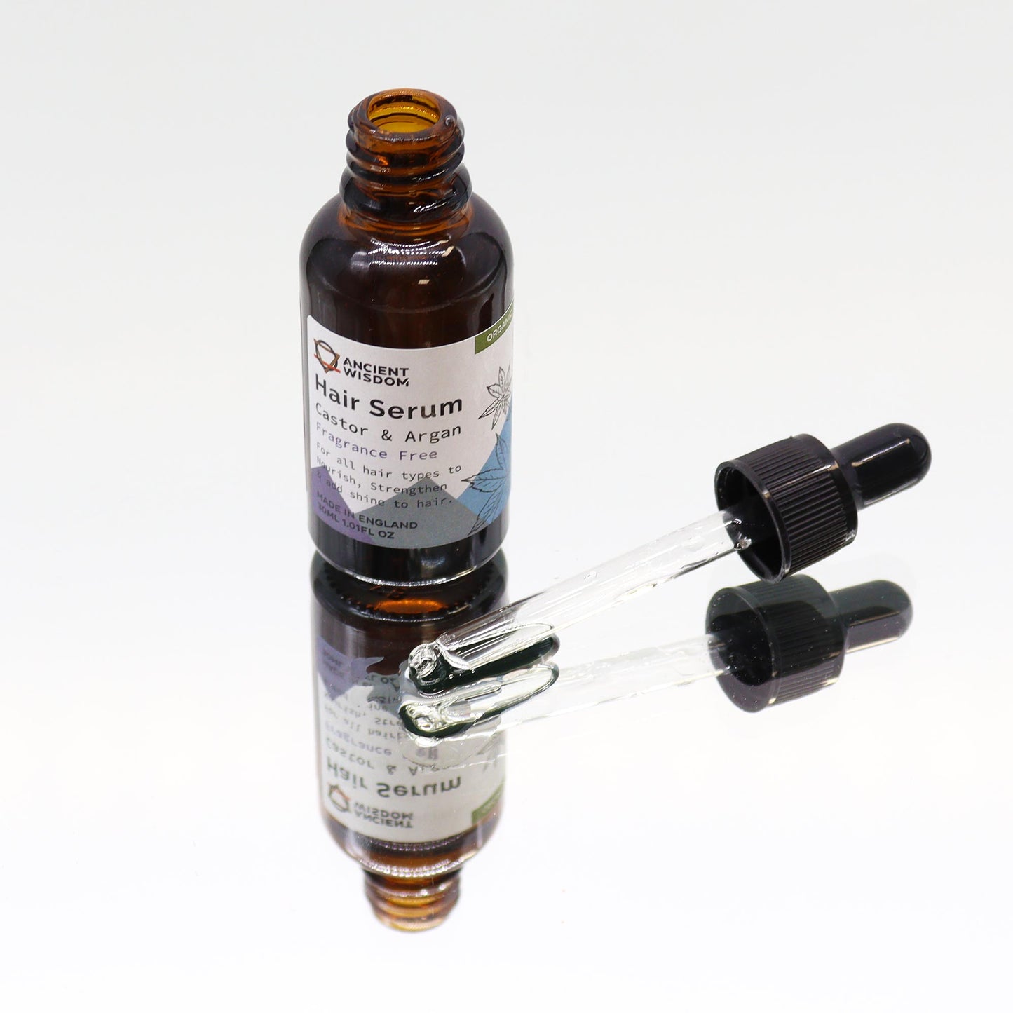 A brown glass bottle of "ANCIENT WISDOM Hair Serum Castor & Argan Fragrance Free" rests on a reflective white surface, with its glass dropper lying to the side.