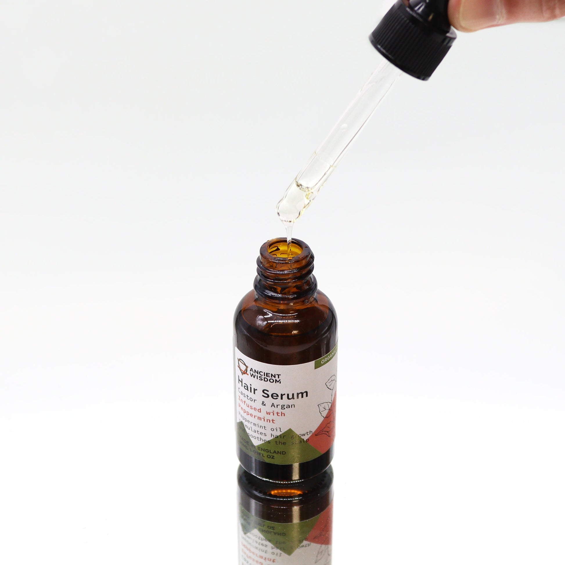 A bottle of "Ancient Wisdom Hair Serum Castor & Argan infused with Peppermint" sits on a white surface as a dropper drips serum into it.