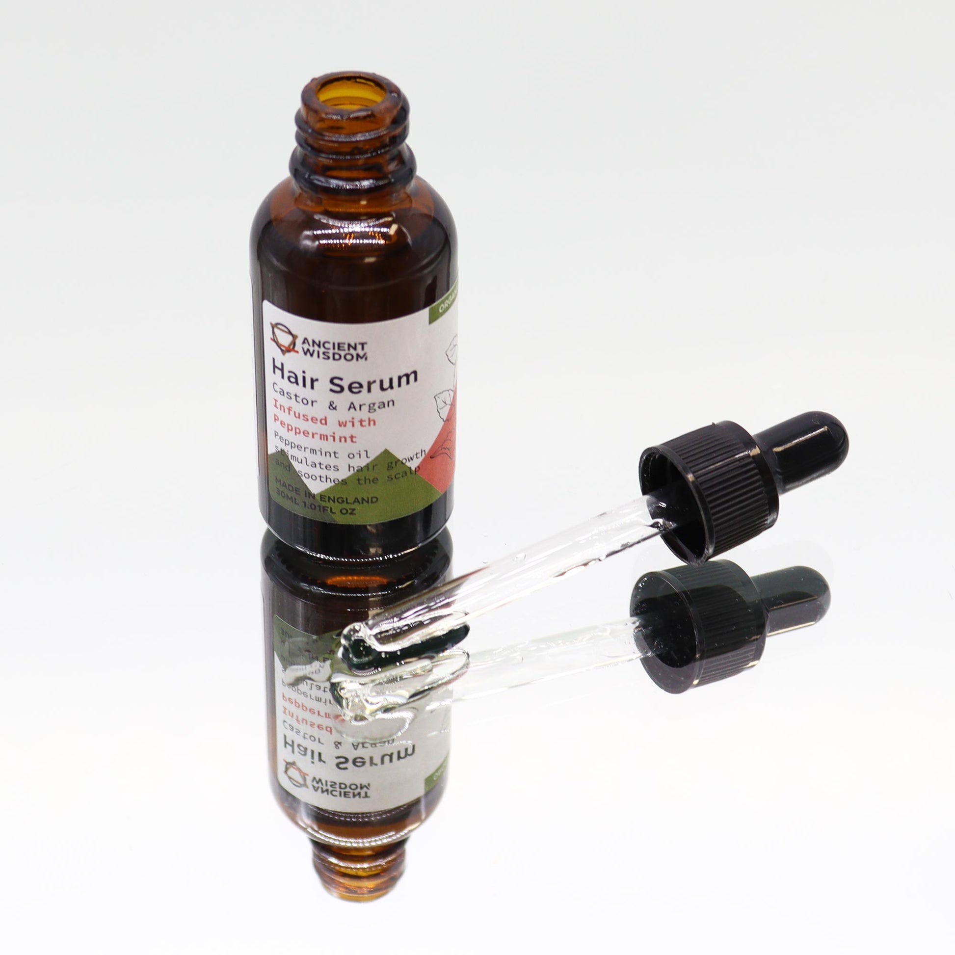 A brown bottle of Ancient Wisdom Hair Serum with its label reading “Hair Serum, Castor & Argan, Infused with Peppermint” sits on a white, reflective surface. A dropper cap lays detached next to the bottle.