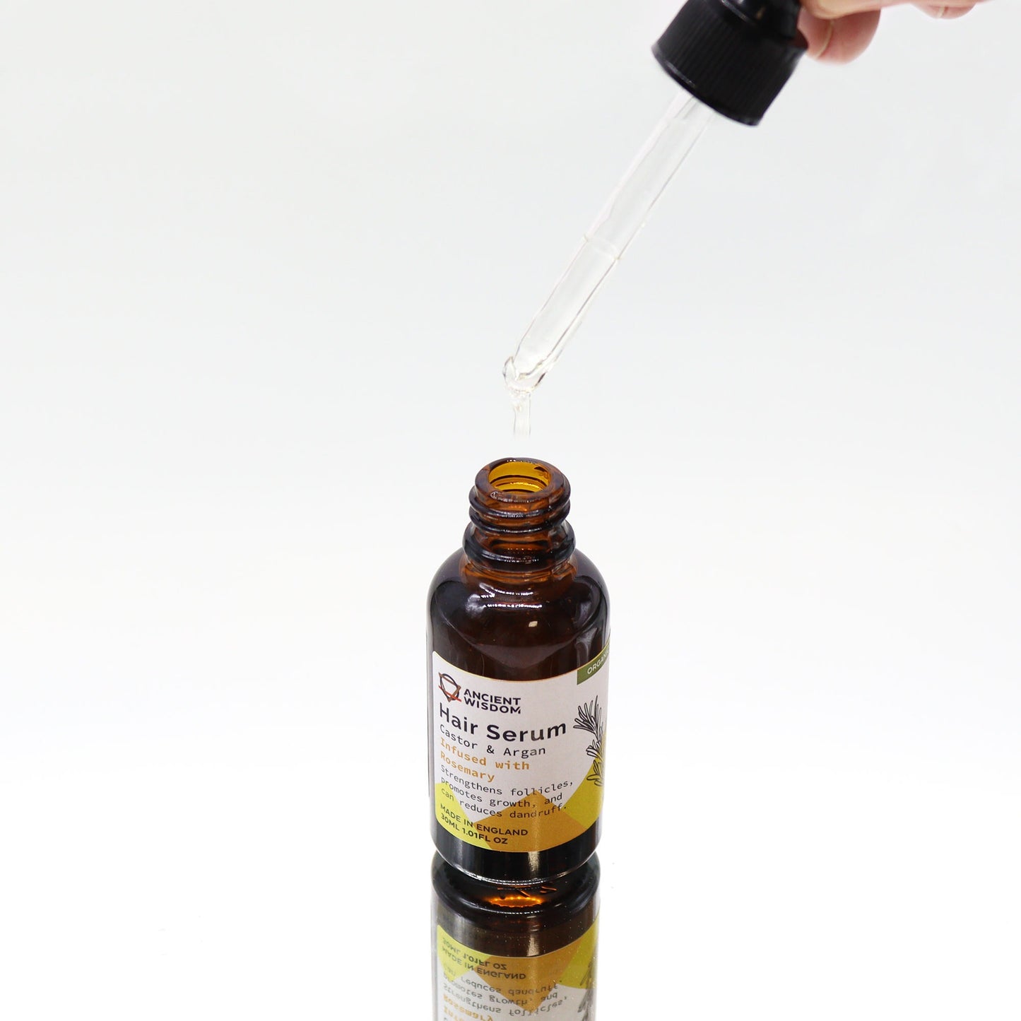 A hand holds a dropper, dispensing clear serum into an open brown "Ancient Wisdom Hair Serum" bottle on a white surface. Bottle text: "Castor & Argan infused with Rosemary. Strengthens follicles... reduces dandruff. Made in England 30ML 1.01FL OZ"