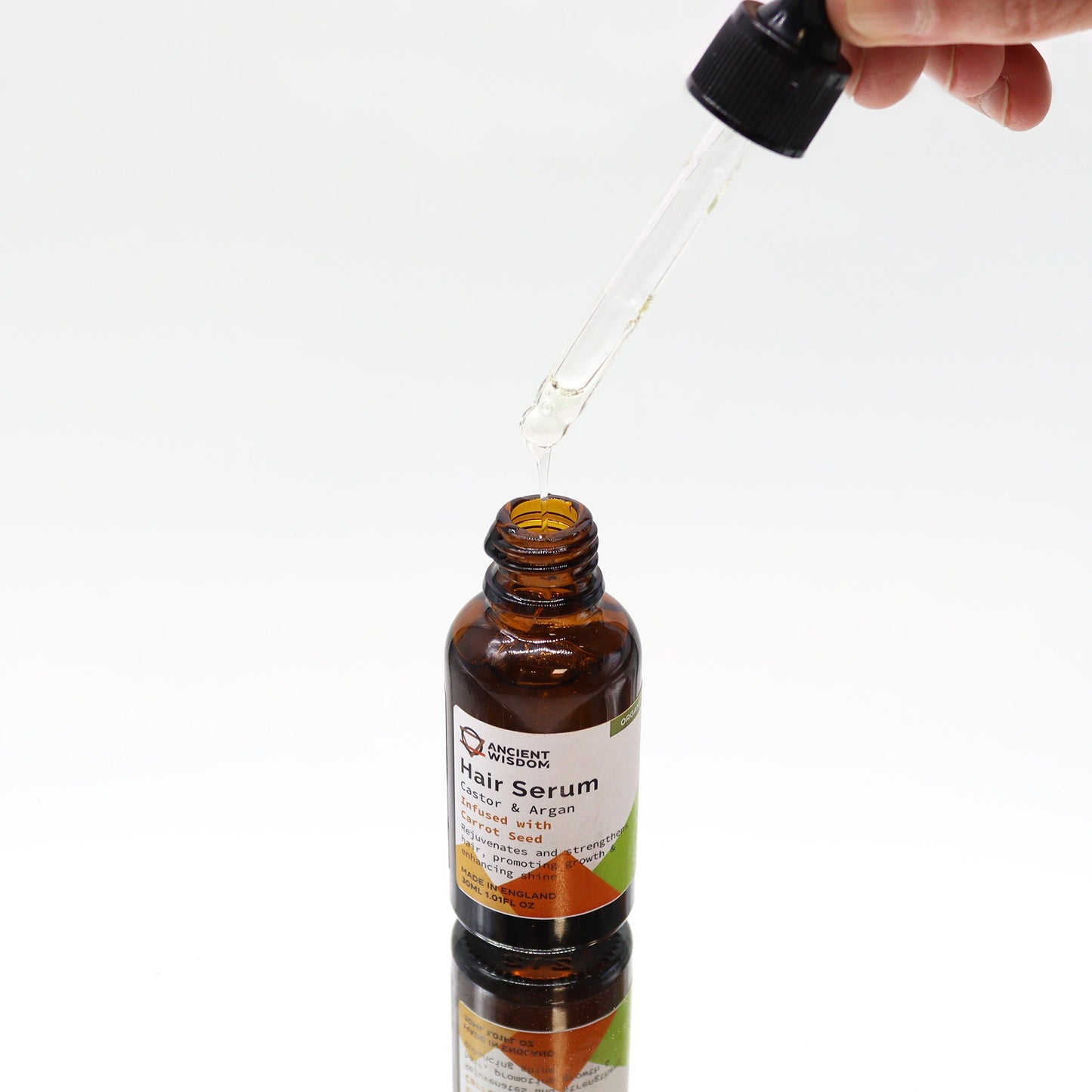 A dropper is dispensing "Ancient Wisdom Hair Serum: Castor & Argan Infused with Carrot Seed" into the amber bottle on a white surface.