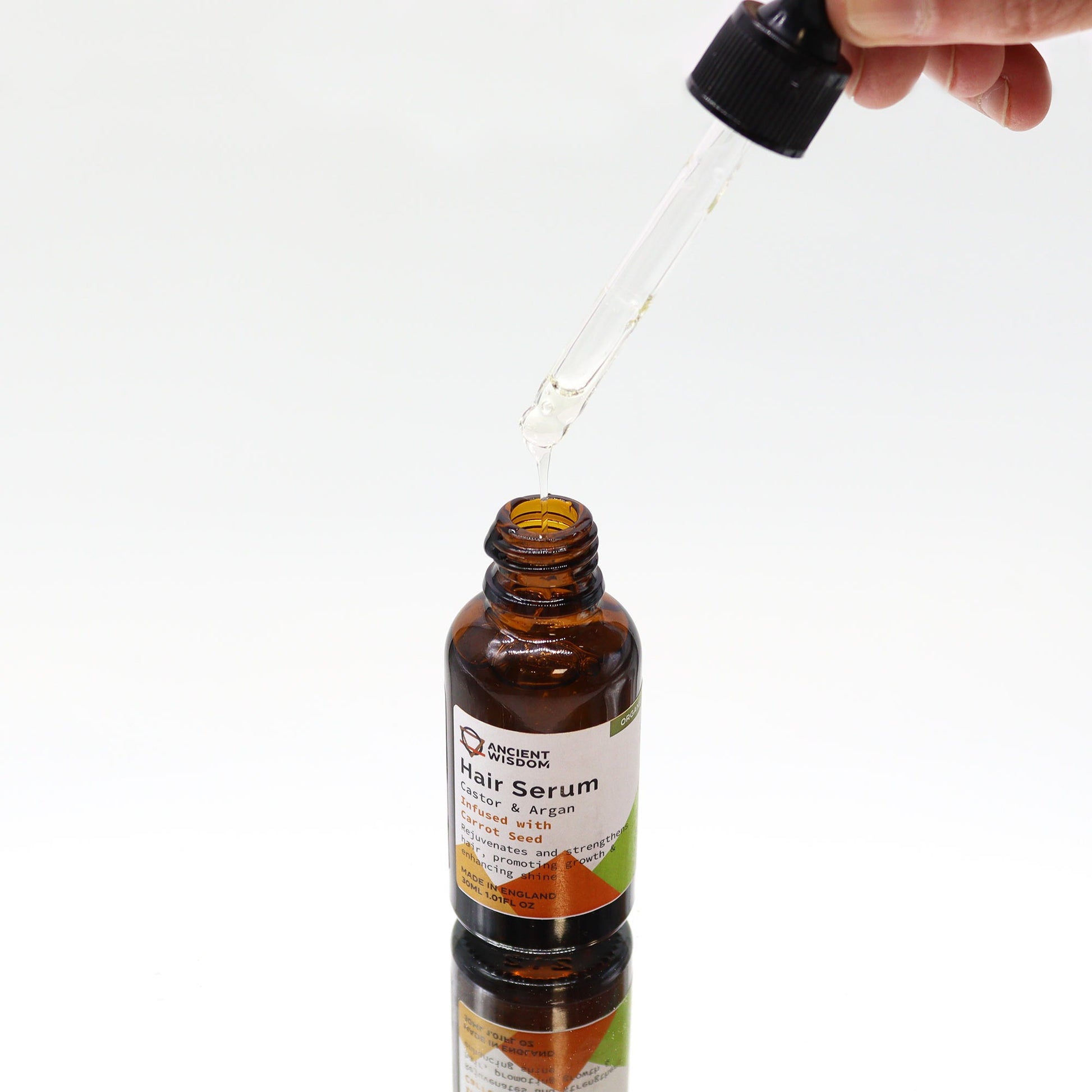 A dropper is dispensing "Ancient Wisdom Hair Serum: Castor & Argan Infused with Carrot Seed" into the amber bottle on a white surface.