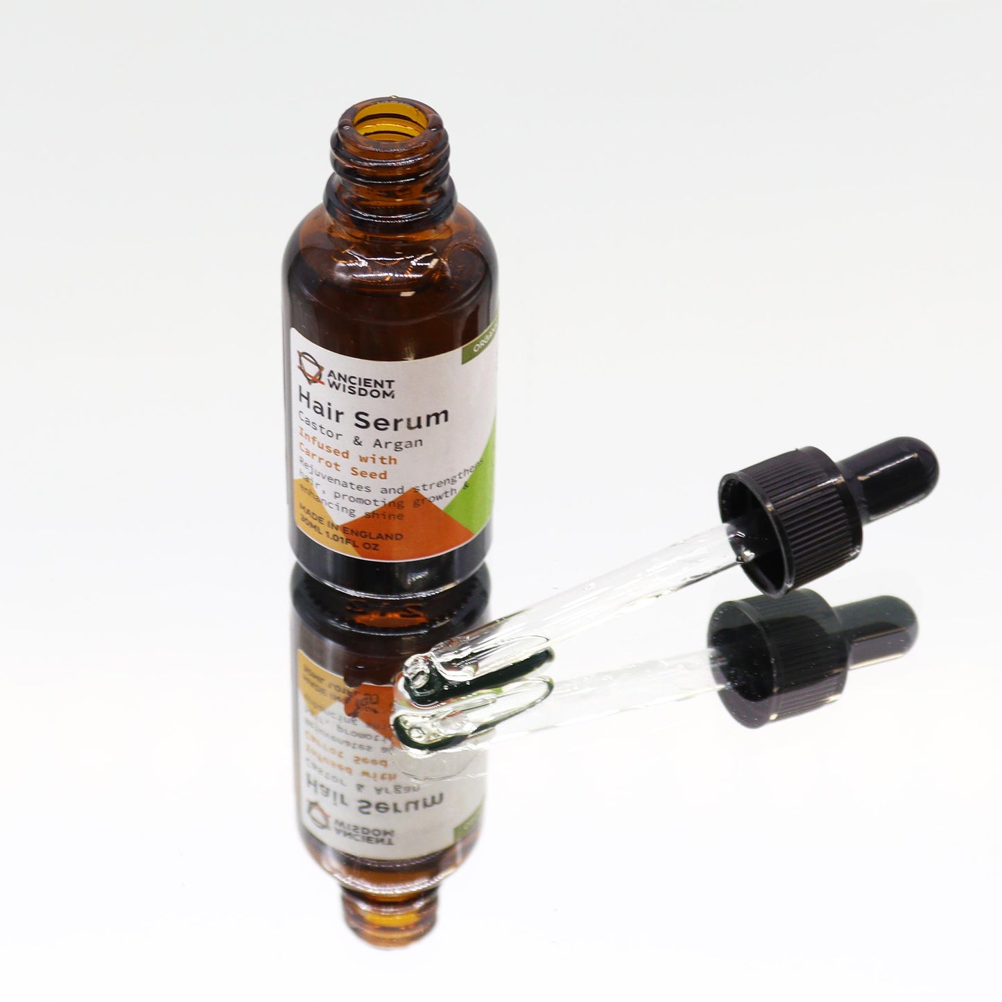 A brown bottle labeled “ANCIENT WISDOM Hair Serum Castor & Argan Infused with Carrot Seed…MADE IN ENGLAND 30ML 1.01FL OZ.” stands on a mirrored surface with its dropper removed, lying next to it in a white space.