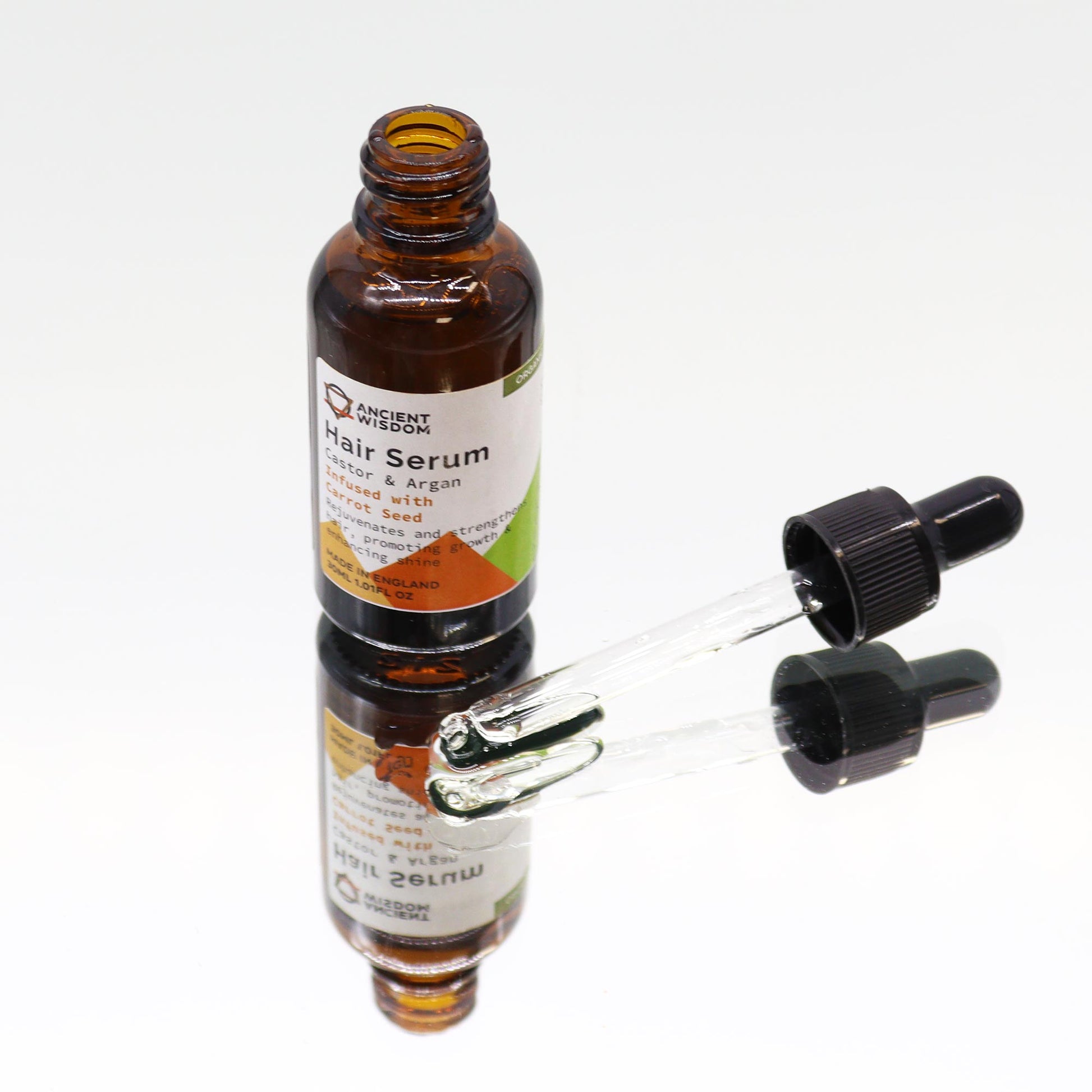 A brown bottle labeled “ANCIENT WISDOM Hair Serum Castor & Argan Infused with Carrot Seed…MADE IN ENGLAND 30ML 1.01FL OZ.” stands on a mirrored surface with its dropper removed, lying next to it in a white space.