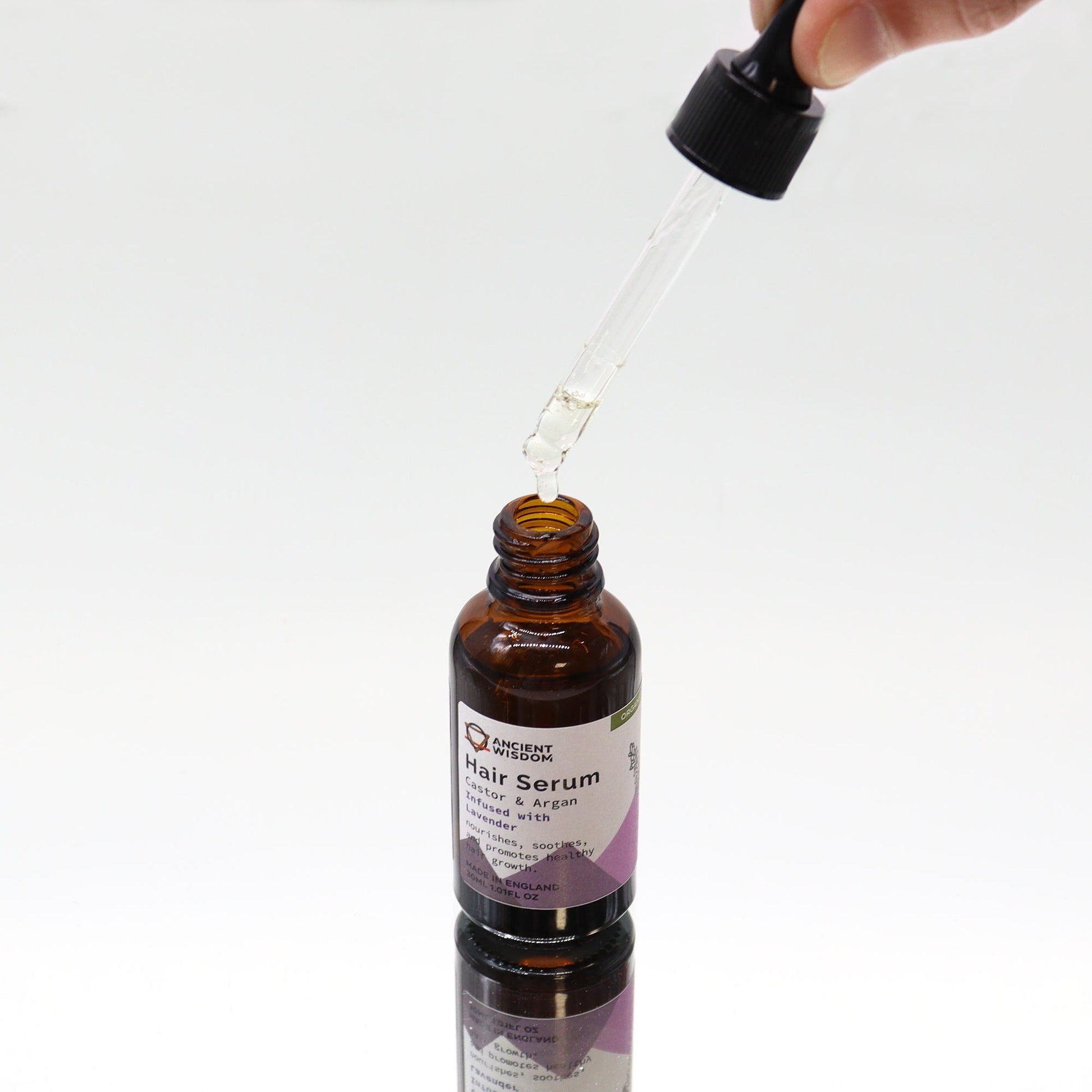 A dropper is dispensing liquid into a small brown glass bottle of "ANCIENT WISDOM Hair Serum Castor & Argan Infused with Lavender, nourishes, soothes, and promotes healthy hair growth. MADE IN ENGLAND 30ML 1.01FL OZ" on a white surface.