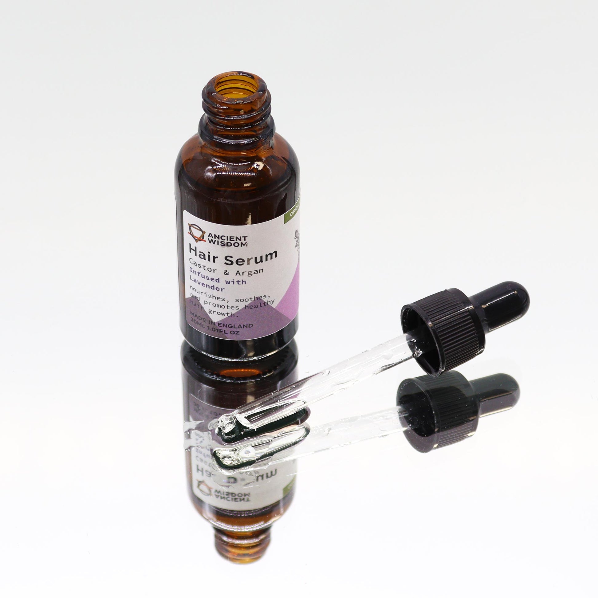A bottle of “ANCIENT WISDOM Hair Serum: Castor & Argan Infused with Lavender” stands on a reflective surface. An open dropper lays on its side next to the bottle.