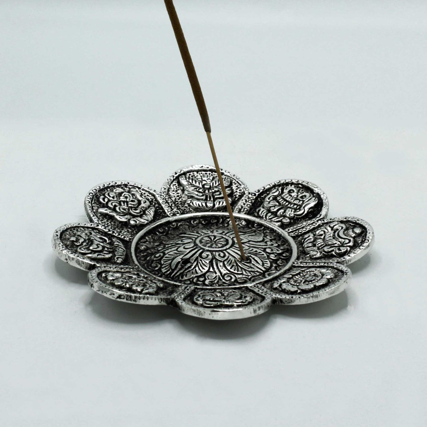 An ornate, circular metal incense holder with floral and animal motifs is holding a single incense stick upright. The setting is a plain, light-colored surface.