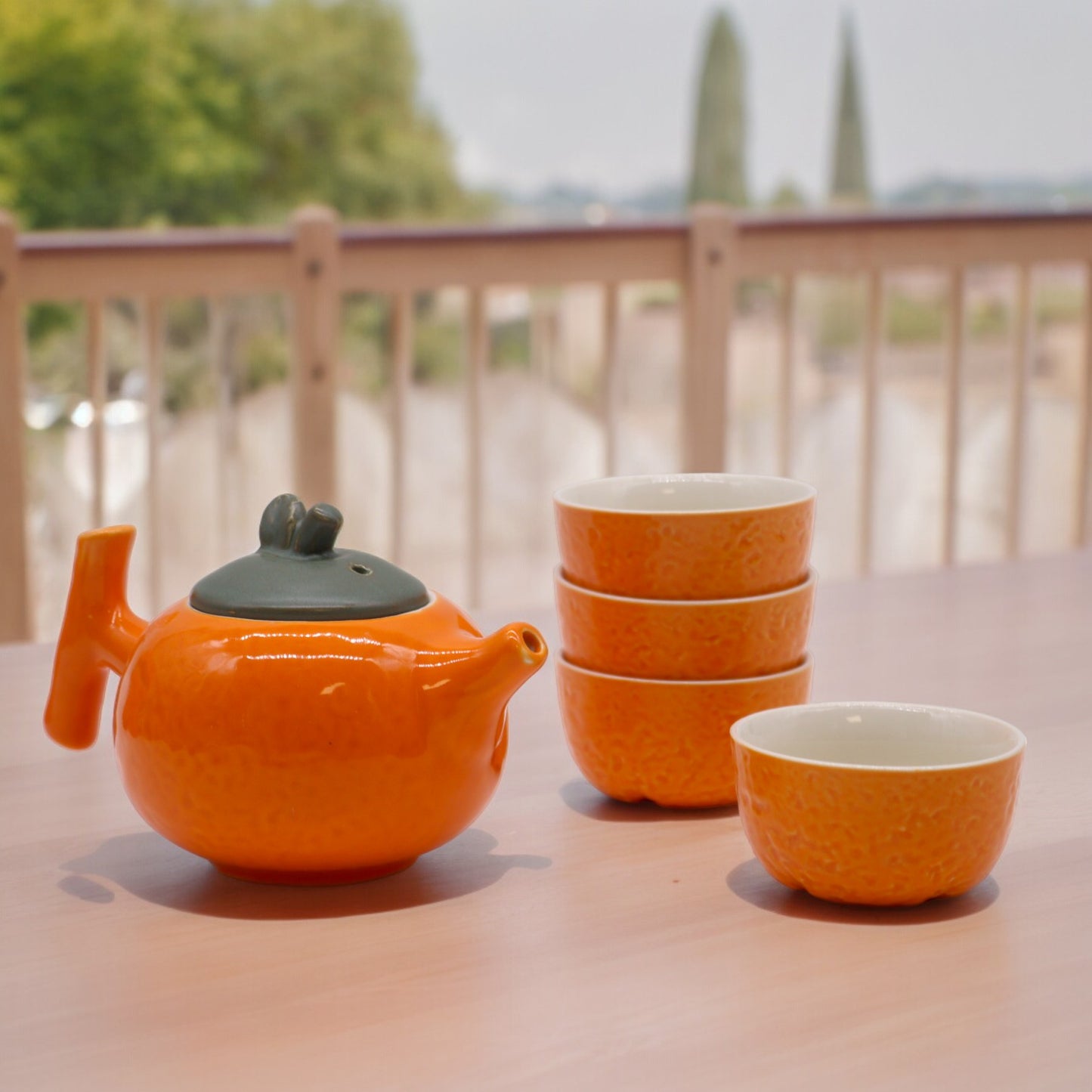 An orange teapot sits on a light wooden table, next to a stack of three and a single orange teacup. The scene is outdoors, with a blurred background of greenery and a wooden fence.