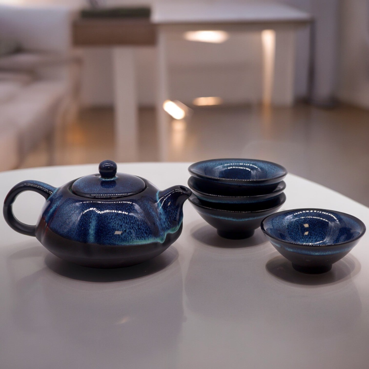 A dark-blue teapot sits on a white table, next to a stack of similar-colored small bowls.  The scene is softly lit, with a blurred background suggesting an interior space.