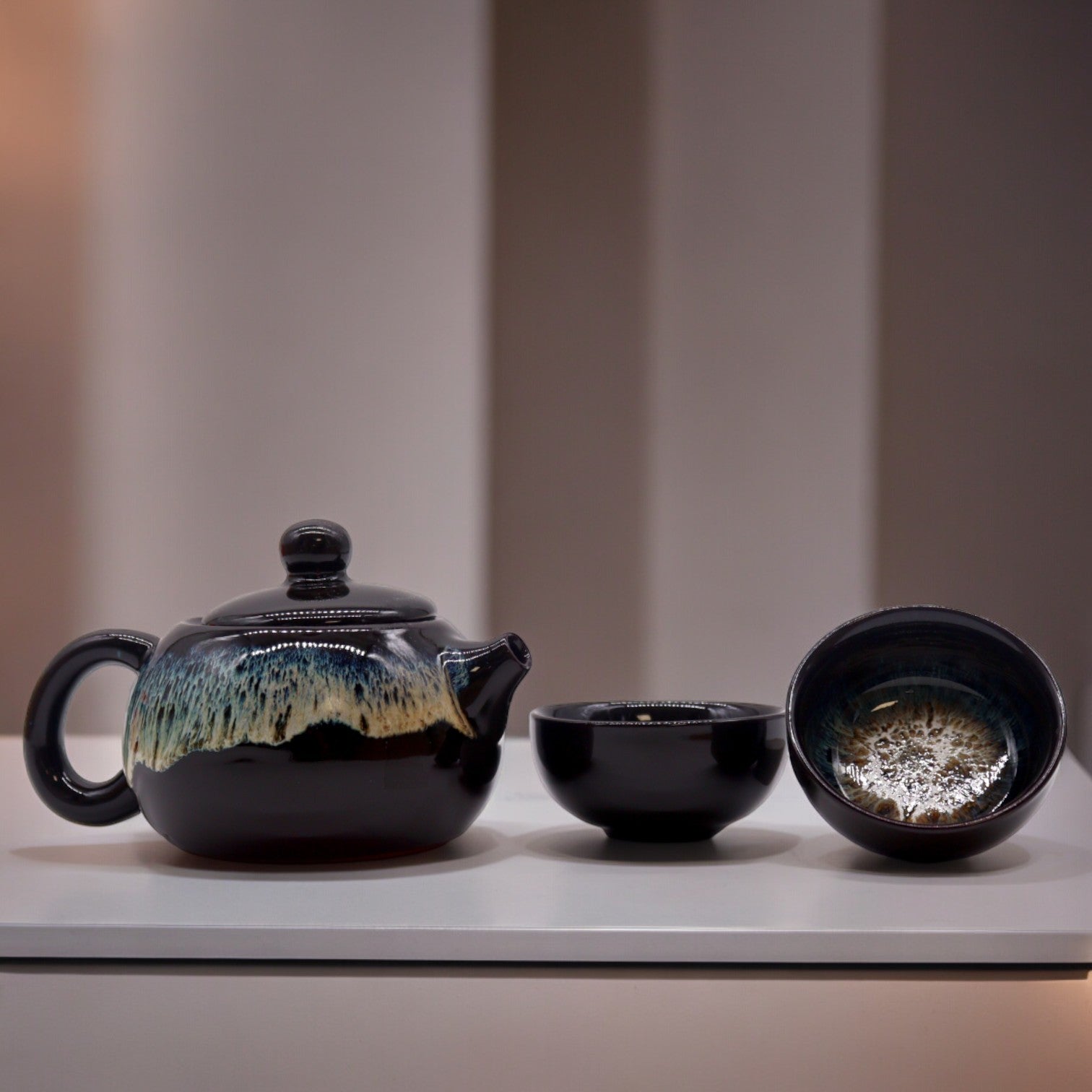 A dark-glazed teapot and two matching bowls sit on a white surface against a blurred background.