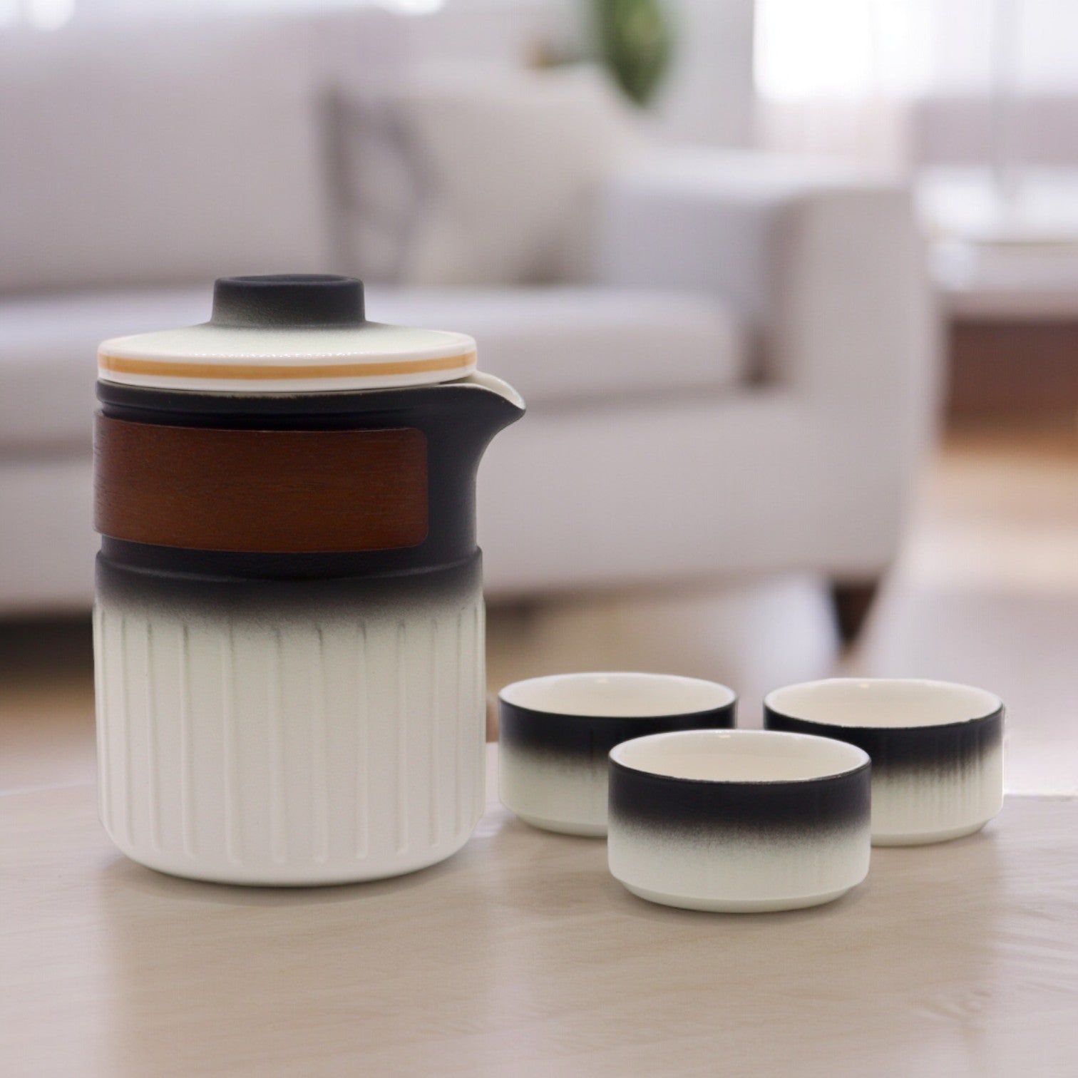 A ceramic teapot sits on a table, accompanied by three small bowls. The teapot features a dark top, a wooden band, and a white, ribbed body; it rests on a light wood surface in a blurred living room.