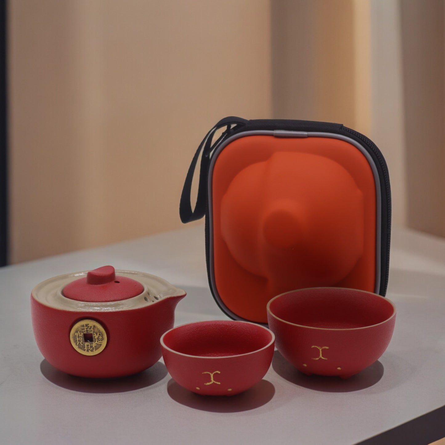 A red ceramic tea set, including a teapot and two cups, sits on a table next to its orange carrying case.