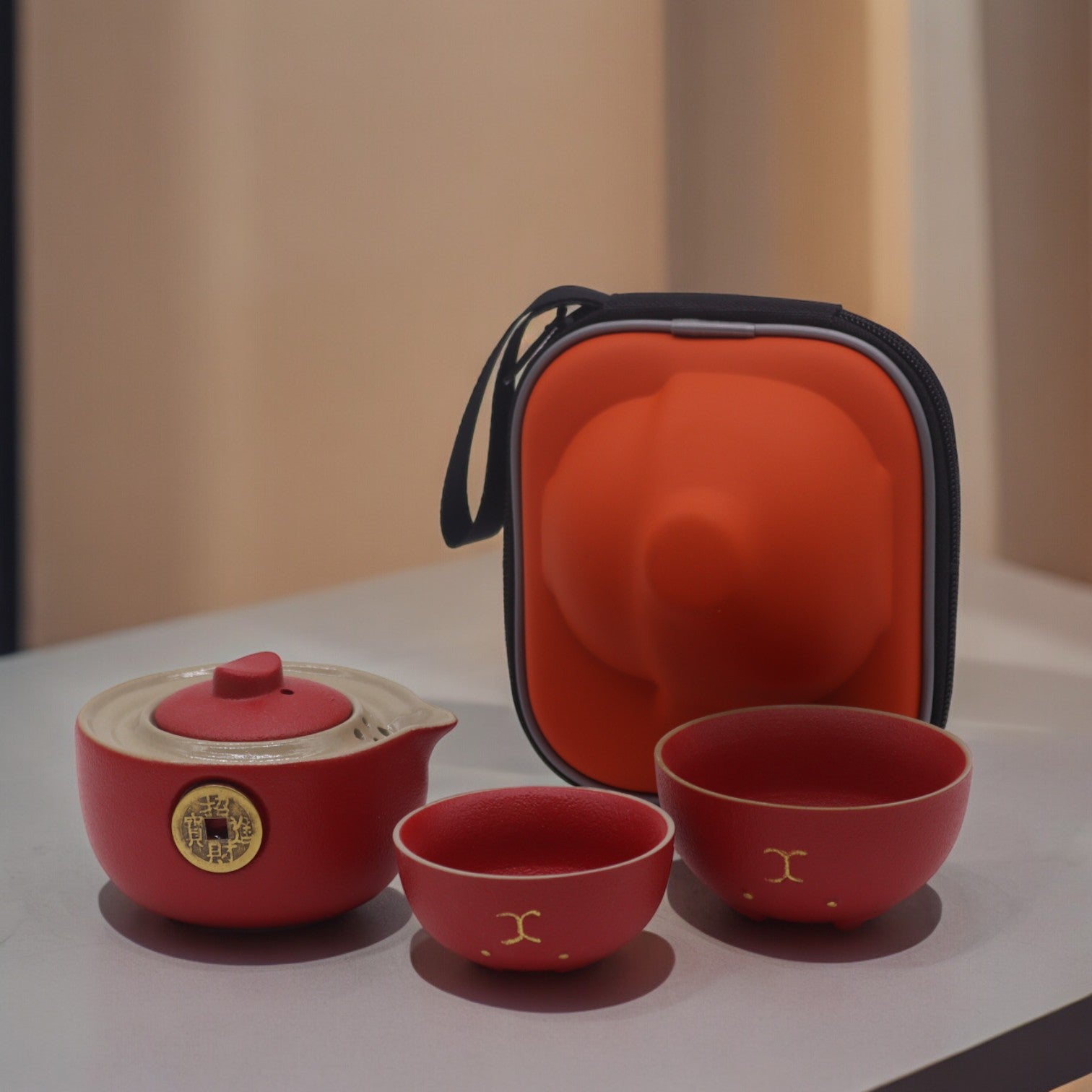 A red ceramic tea set, including a teapot and two cups, sits on a table next to its orange carrying case.