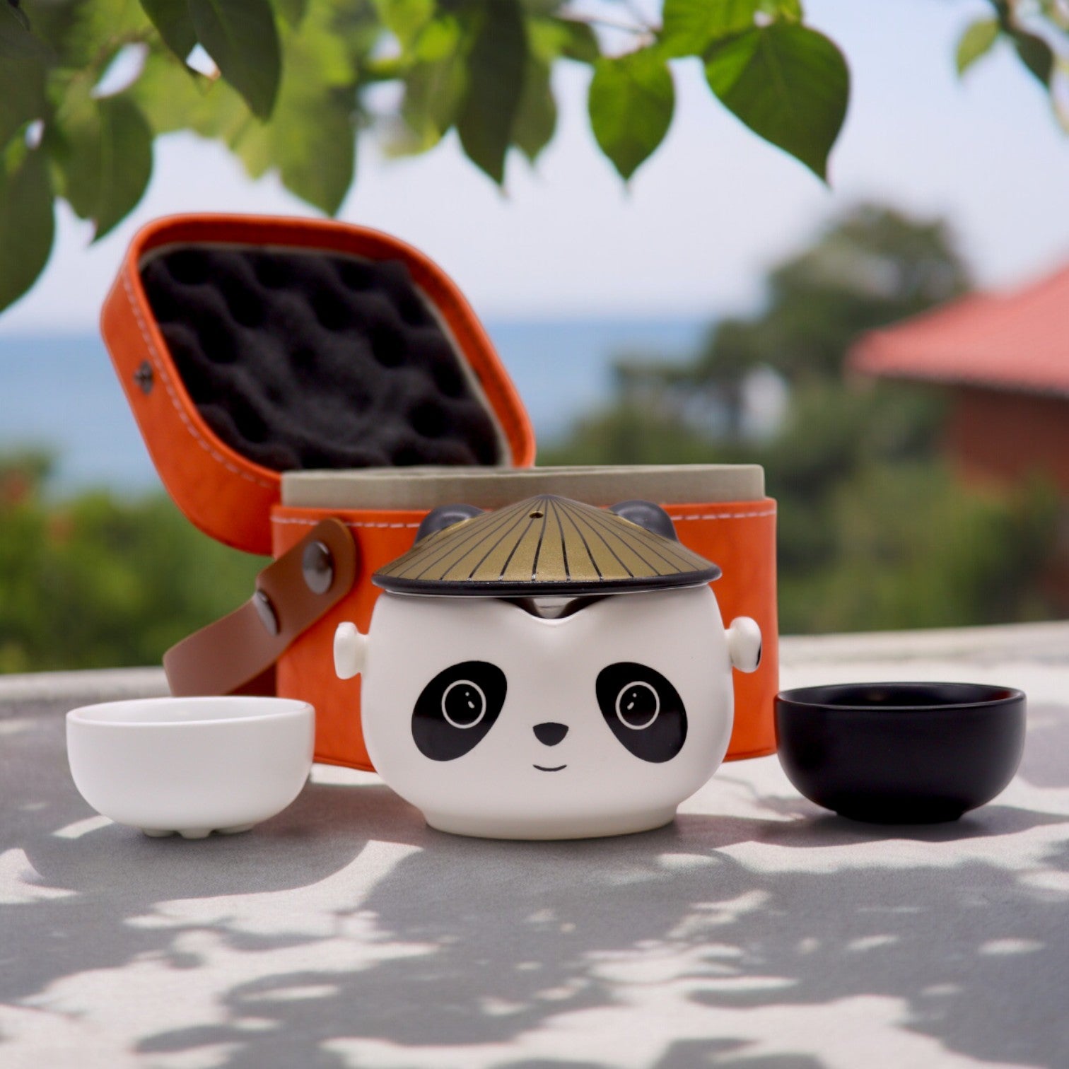 A panda-shaped tea set sits open on a table outdoors.  A small, orange carrying case is partially open behind it. Two small bowls flank the teapot.