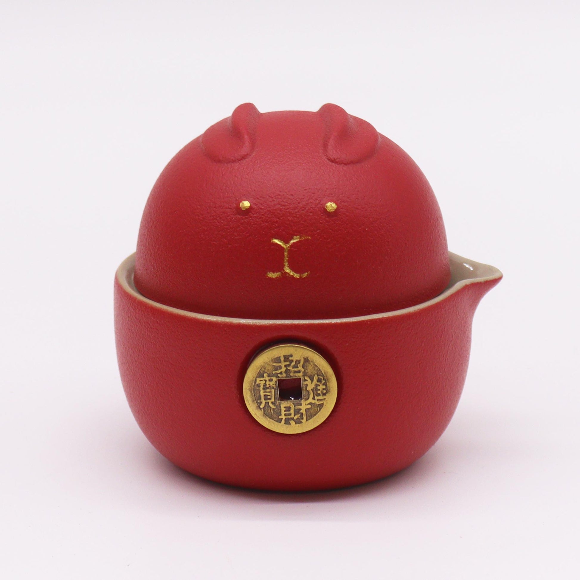 A red ceramic teapot, shaped like a rabbit, sits on a white surface.  The teapot features gold detailing and a brass coin with Chinese characters.