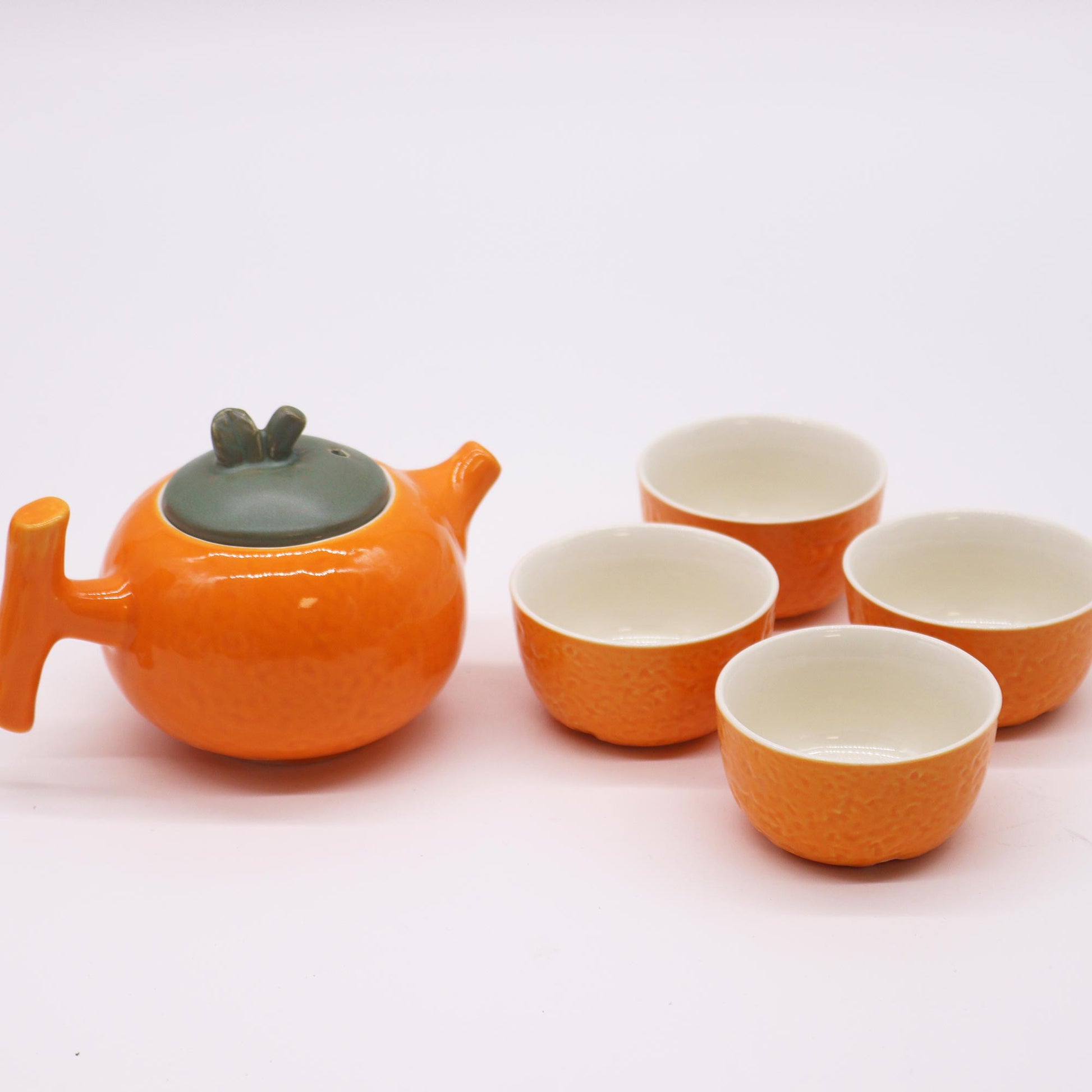 An orange teapot sits with four matching orange cups on a white background.  The teapot and cups appear to be ceramic.