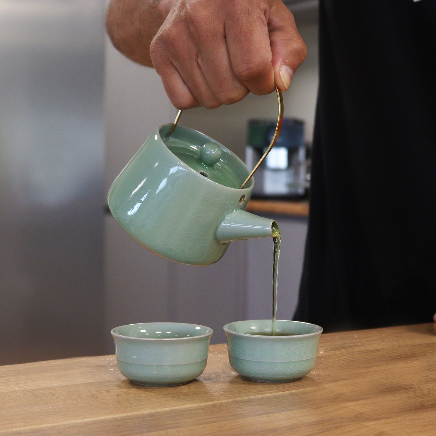 A light-green ceramic teapot pours tea into two matching teacups on a wooden table.