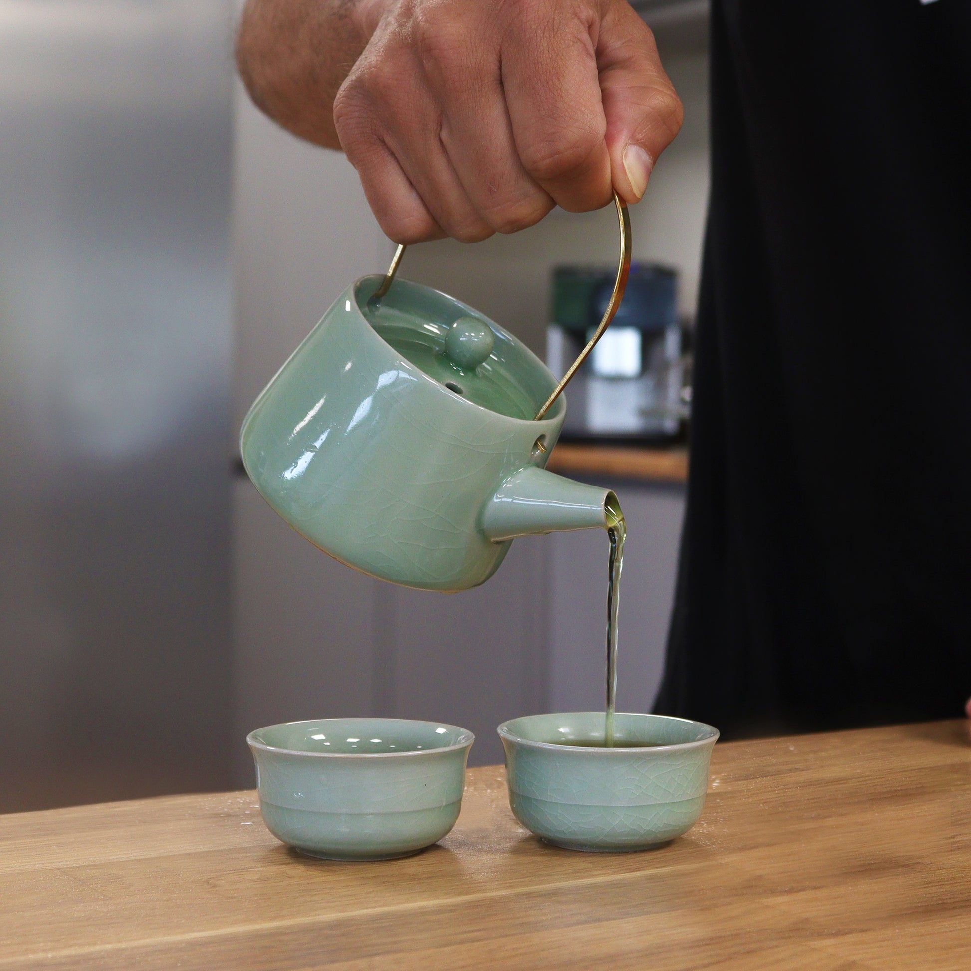 A light-green ceramic teapot pours tea into two matching teacups on a wooden table.