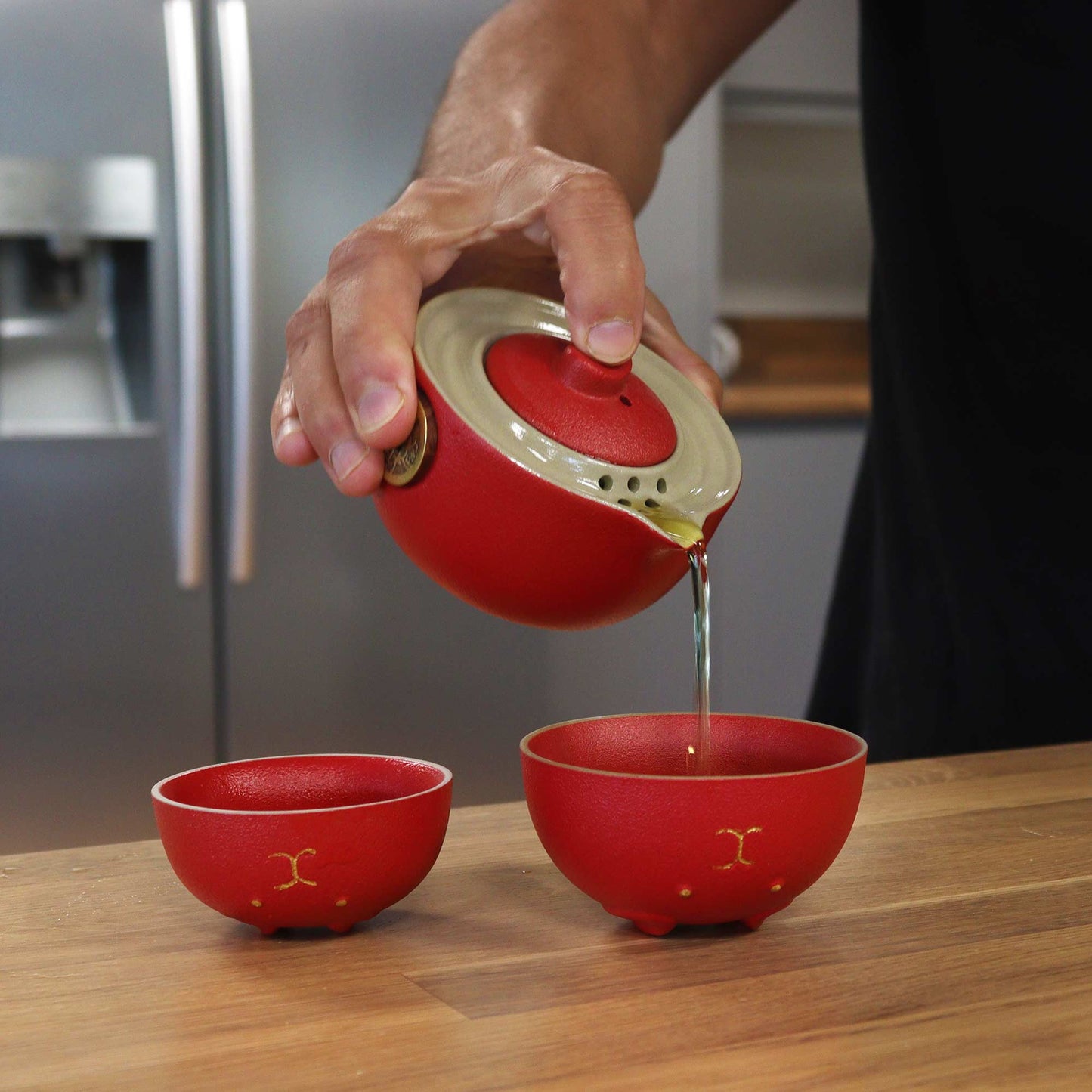 A red teapot pours liquid into red cups; a person's hand holds the teapot; the setting is a kitchen countertop.