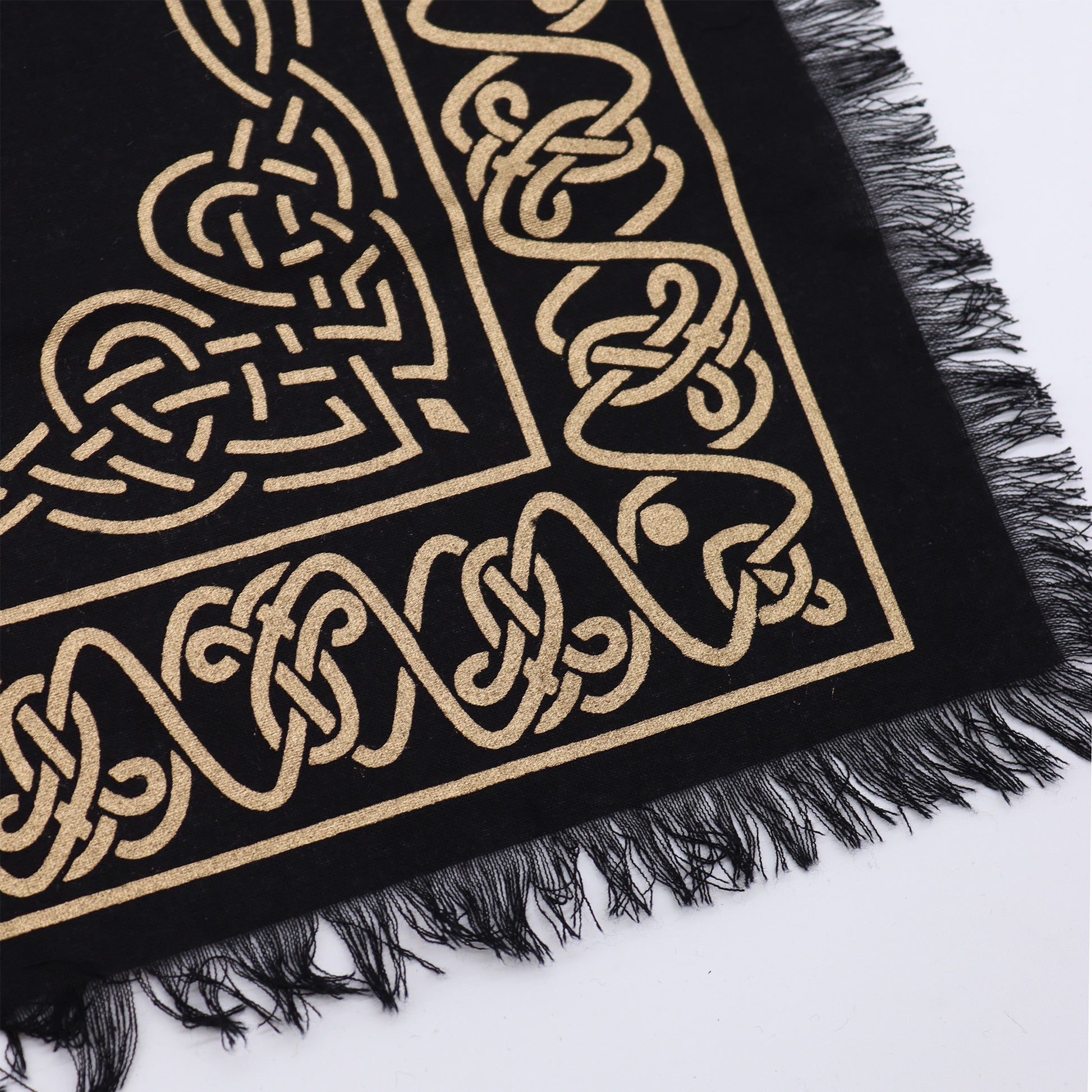 A black fabric square with a gold Celtic knot design is displayed against a white background.  The fabric has a fringed edge.