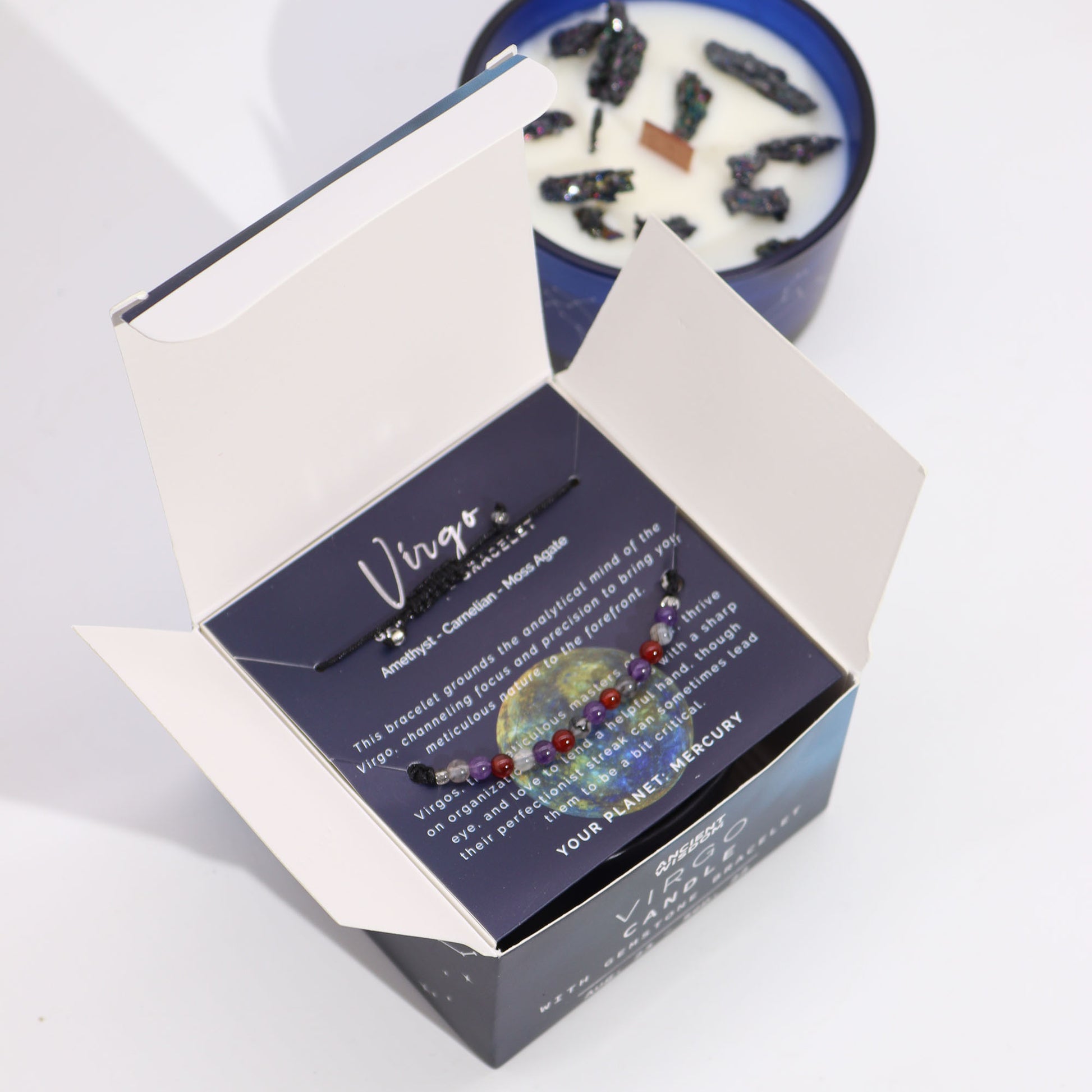 Box contains a Virgo-themed bracelet with amethyst, carnelian, and moss agate on a card. Text: "Virgo...YOUR PLANET: MERCURY." Nearby is a candle with dark crystals embedded in the wax.
