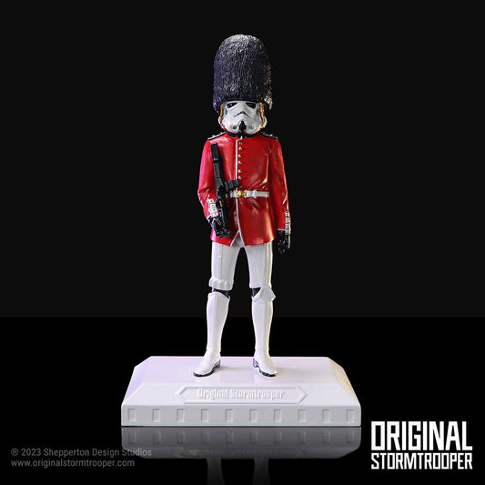Stormtrooper figure wearing a red British guard coat and tall bearskin hat stands at attention on a white display base against a black background; text: "Original Stormtrooper" on base; "ORIGINAL STORMTROOPER"; "© 2023 Shepperton Design Studios www.originalstormtrooper.com"