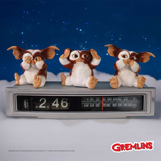 Three Mogwai figurines pose covering eyes, ears, and mouth while sitting on a vintage flip clock radio against a starry blue sky and cloud backdrop; text: "2:46 PM", "FM 88 92 96 100 104 106 MHz", "AM 54 60 70 80 100 120 140 160 KHz", "GREMLINS", "GREMLINS and all related characters and elements © & ™ Warner Bros. Entertainment Inc."