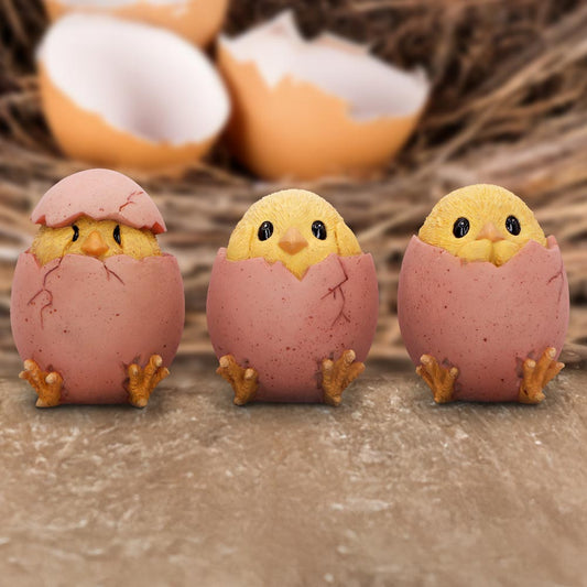 Three yellow chicks peek from cracked pink eggshell halves, sitting in a row on a rough wooden surface with a blurred straw nest and broken eggshells in the background.