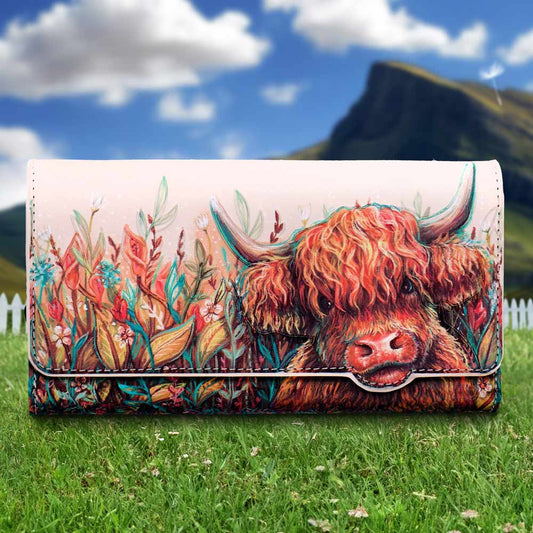 Leather wallet shows a colorful painted Highland cow peeking through wildflowers, placed on green grass before a white picket fence and a distant mountain under a blue sky with clouds.